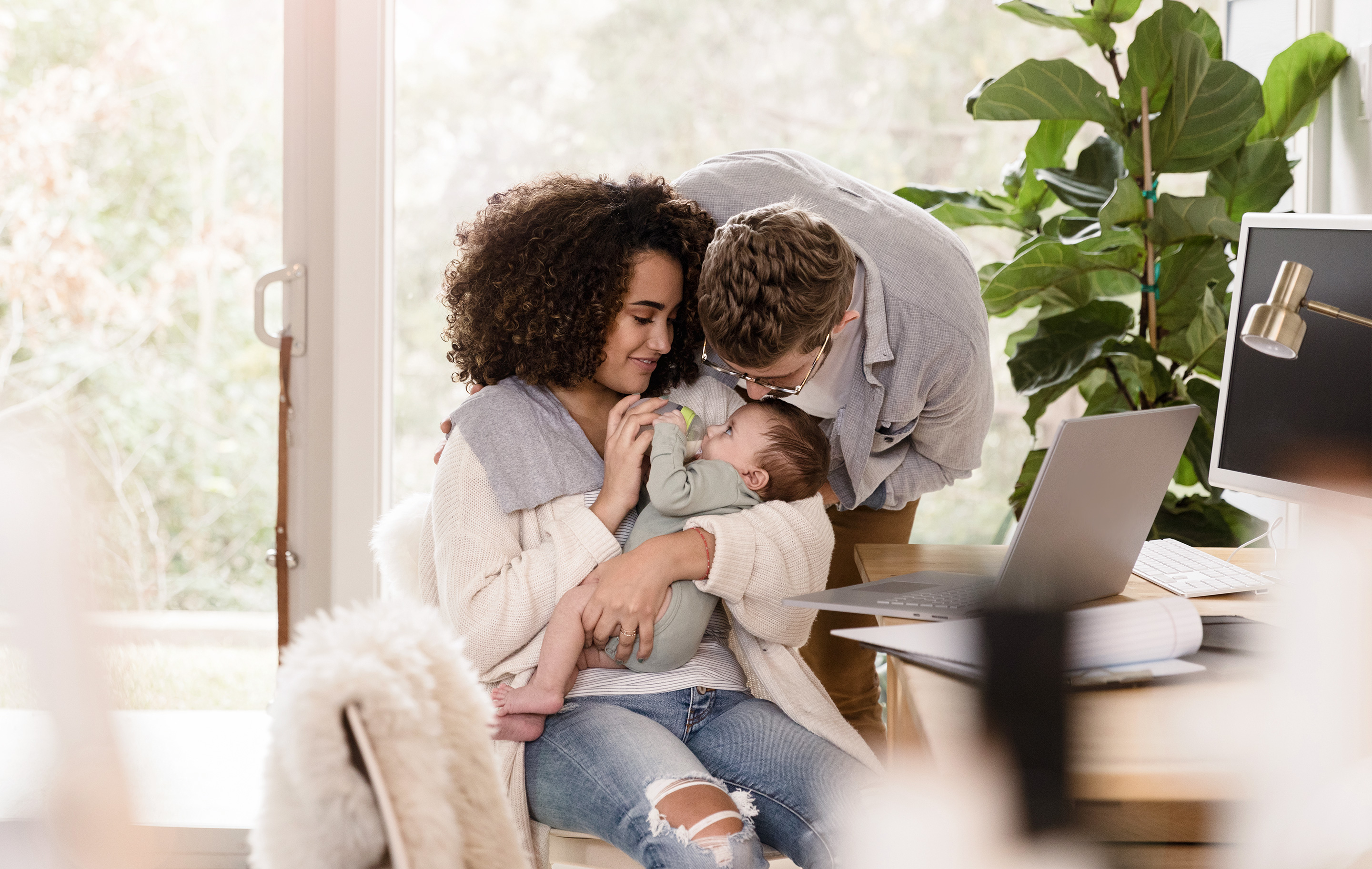 A woman holds a baby while a man kisses its head in a sunlit home office.