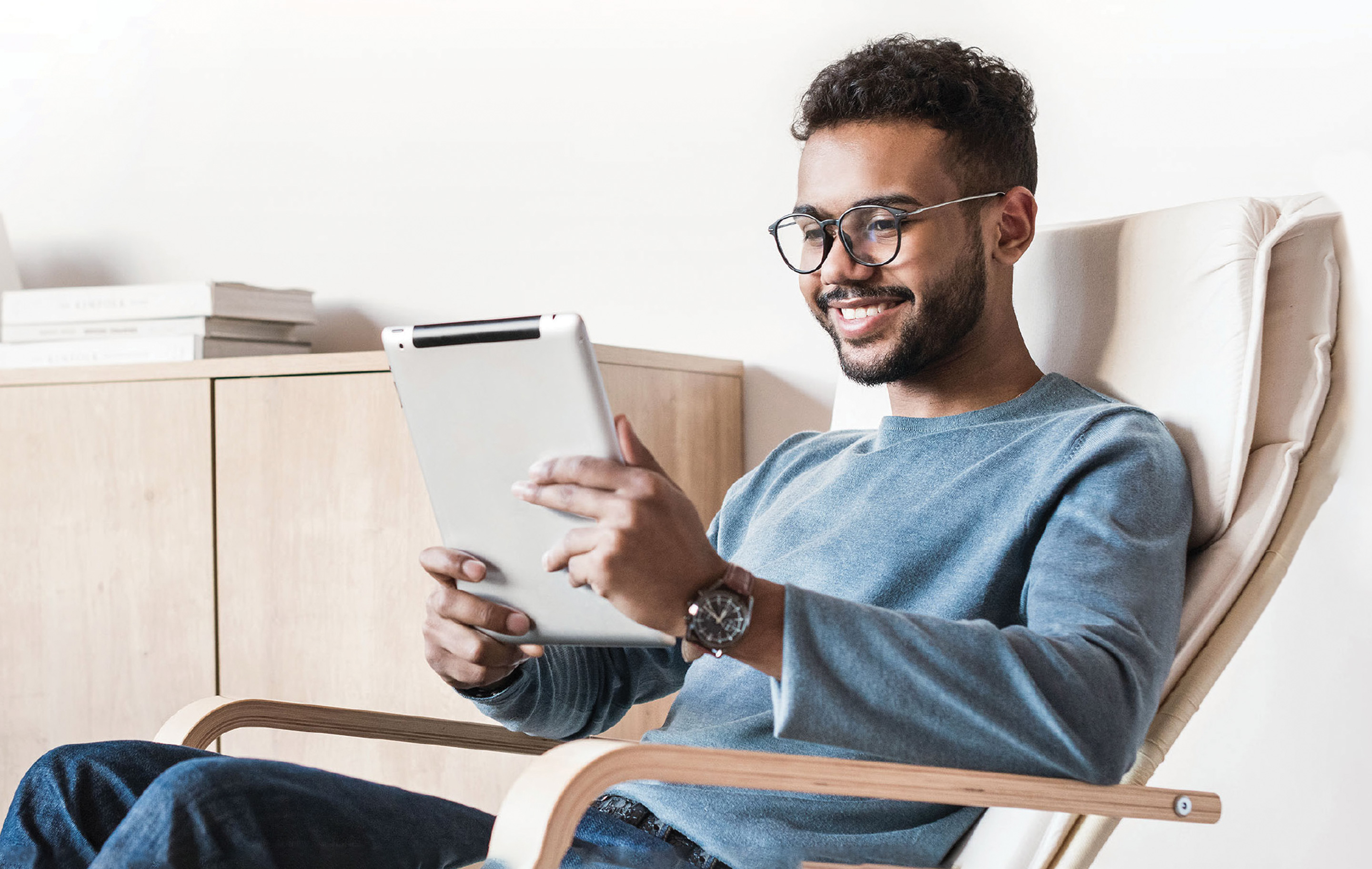 Man with glasses smiles, holding a tablet, seated comfortably in a cozy chair.