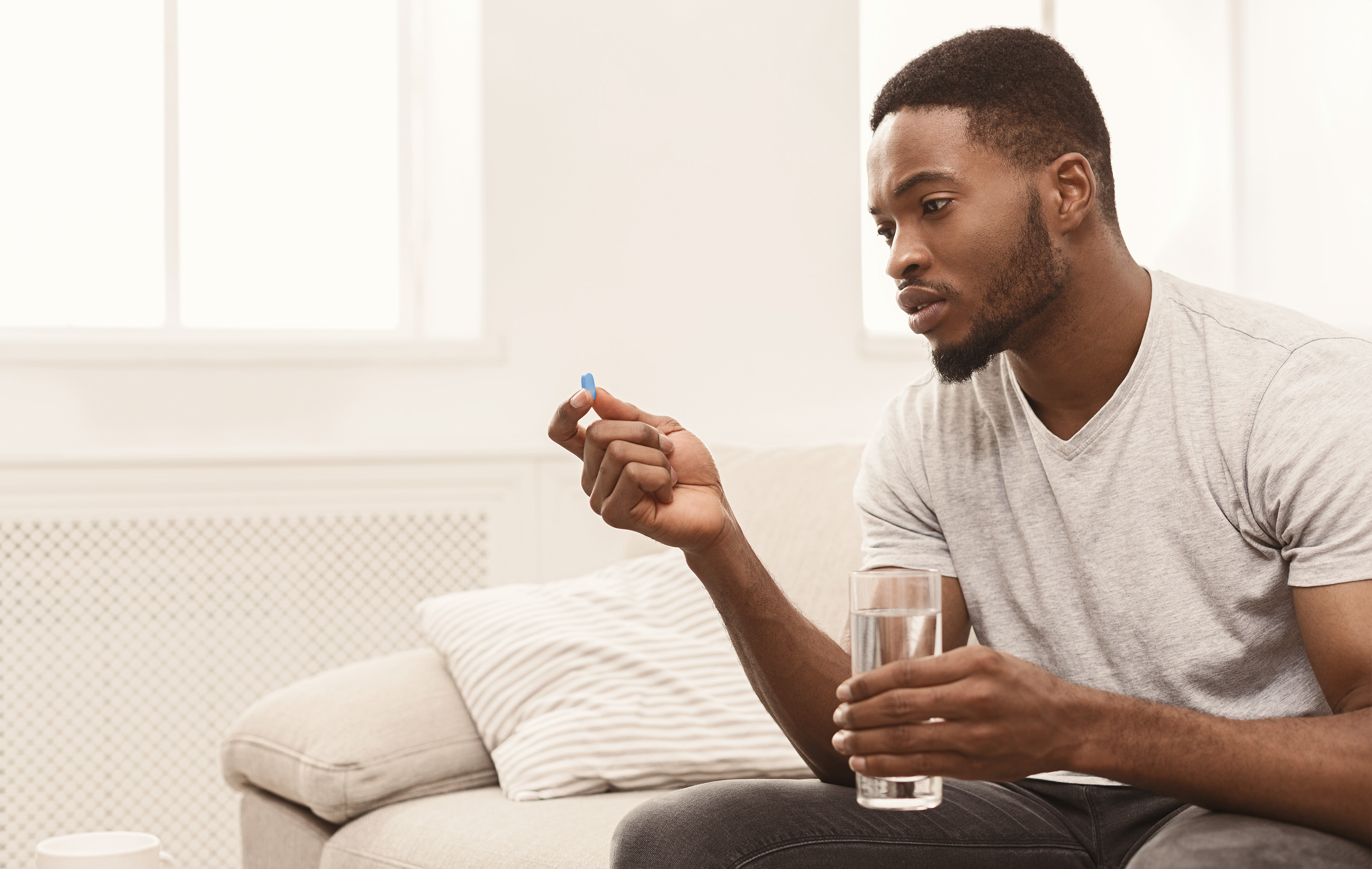 Man sits on couch holding pill and glass of water in bright room