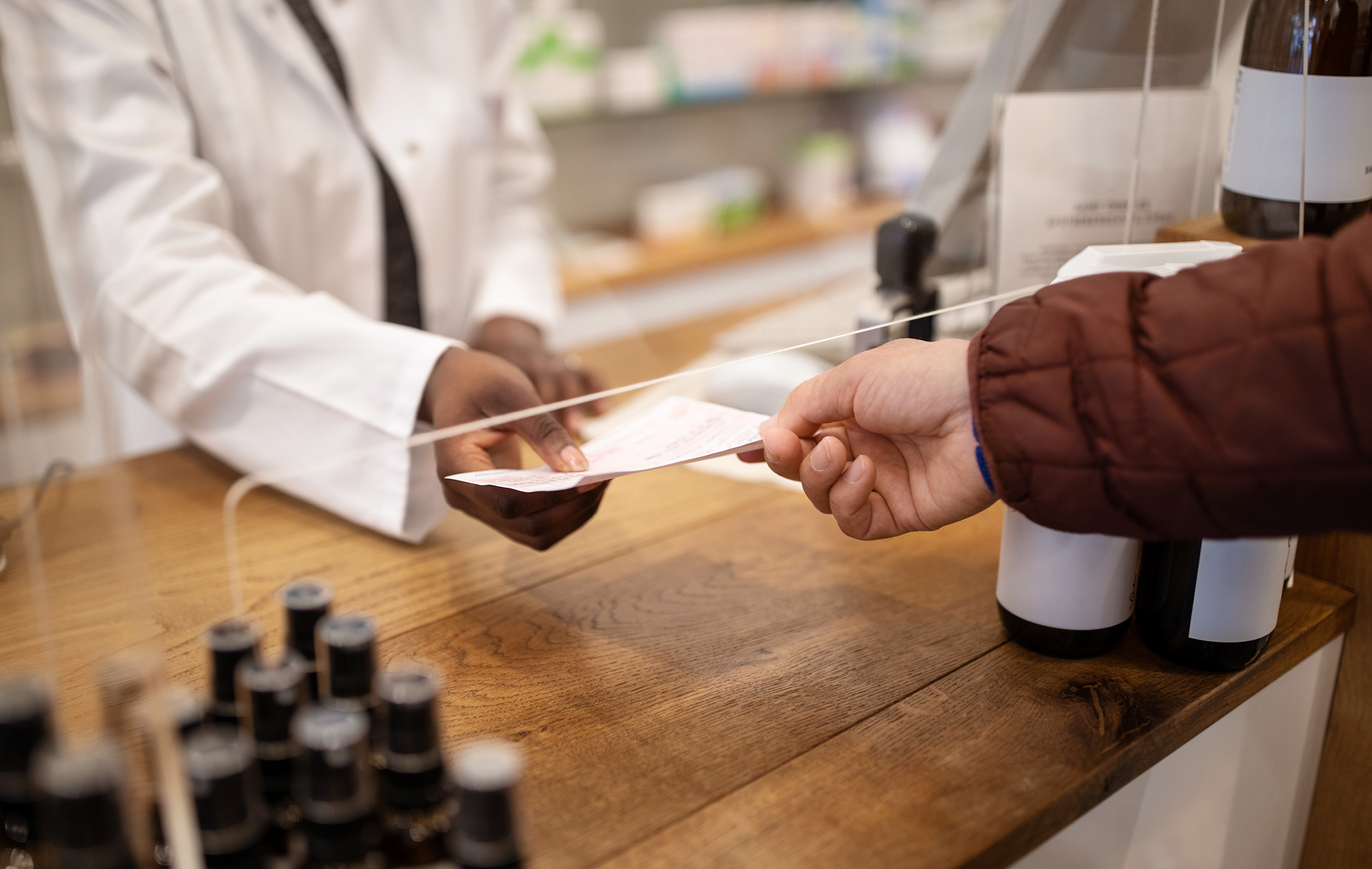 Pharmacist in white coat hands prescription to customer at wooden counter with medicine bottles nearby