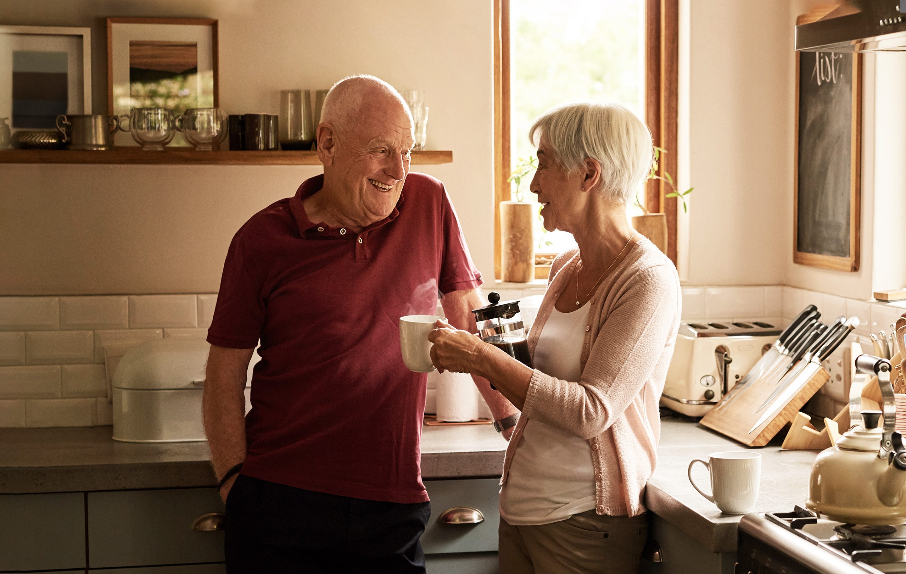 Smiling elderly couple sharing a warm moment in a sunlit kitchen, holding mugs together