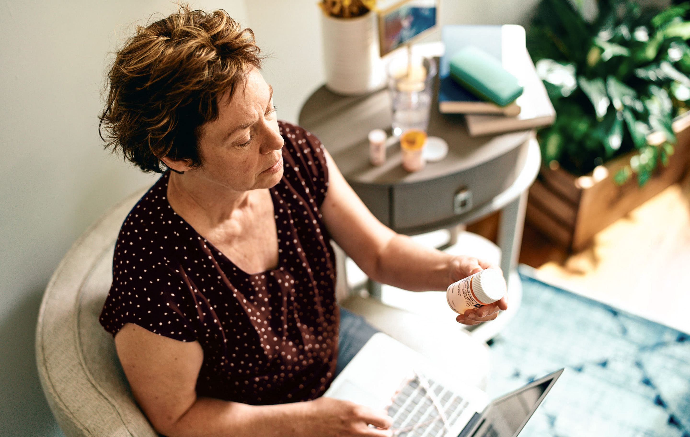 Woman sits in chair, holding pill bottle, works on laptop, sunlight streams through window