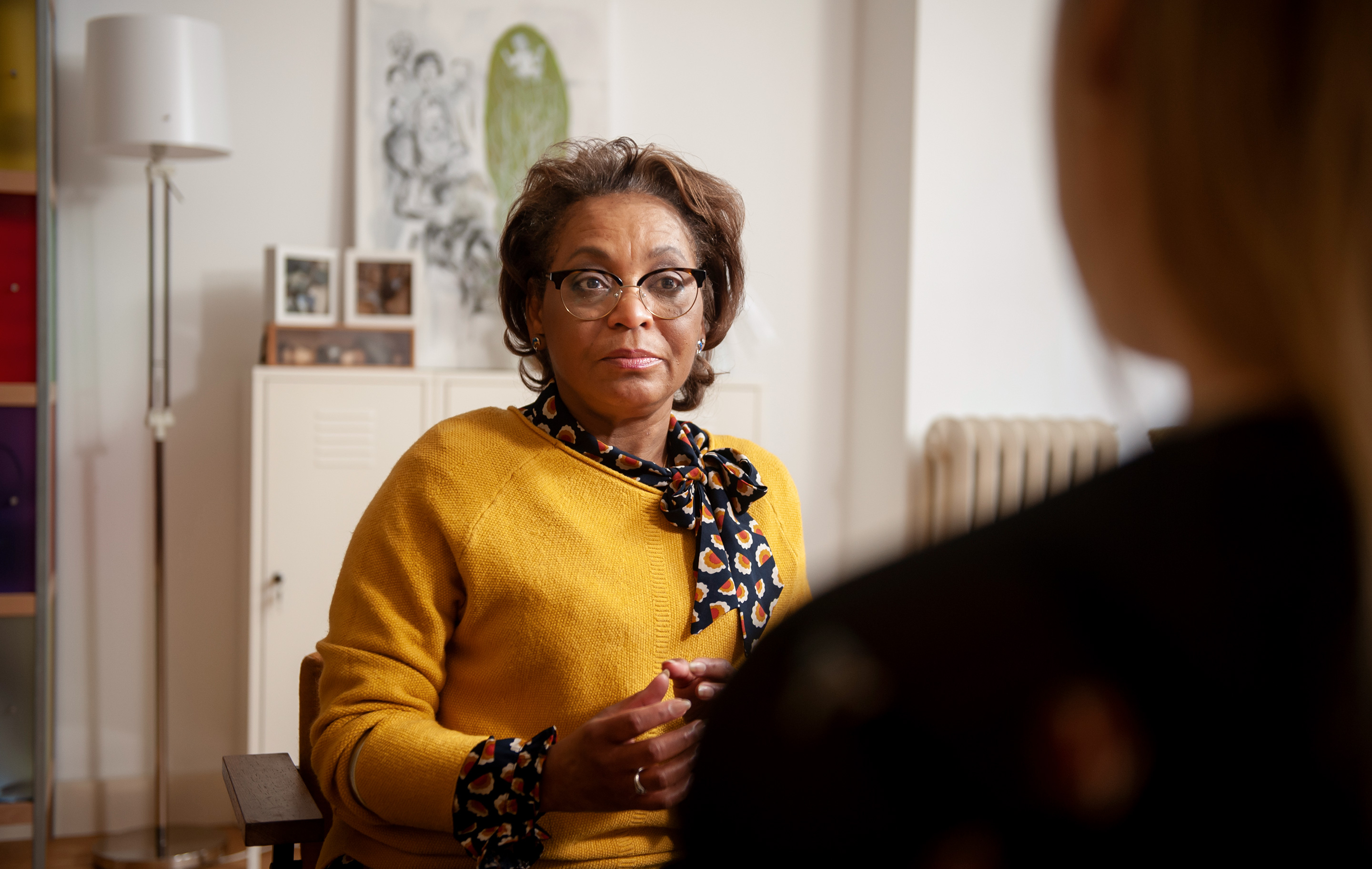 A woman in a yellow sweater and glasses sits facing another person in a warmly lit room.