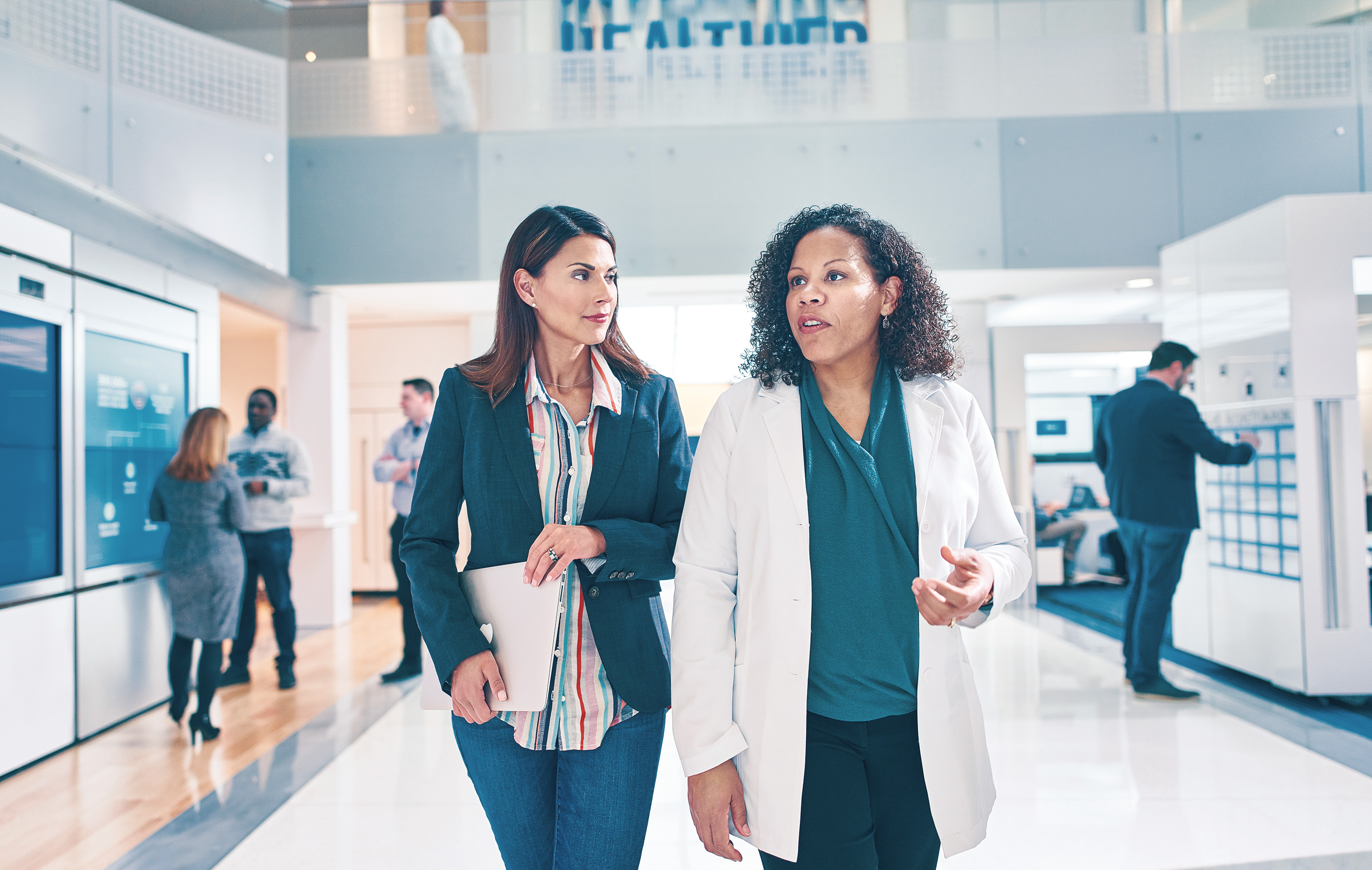 Two women in business attire converse in a bright modern health facility hallway