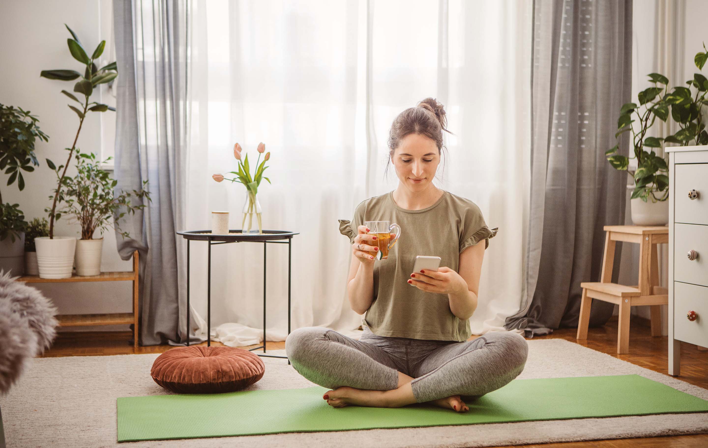 Woman meditates on green mat in bright, plant-filled room holding phone and tea cup