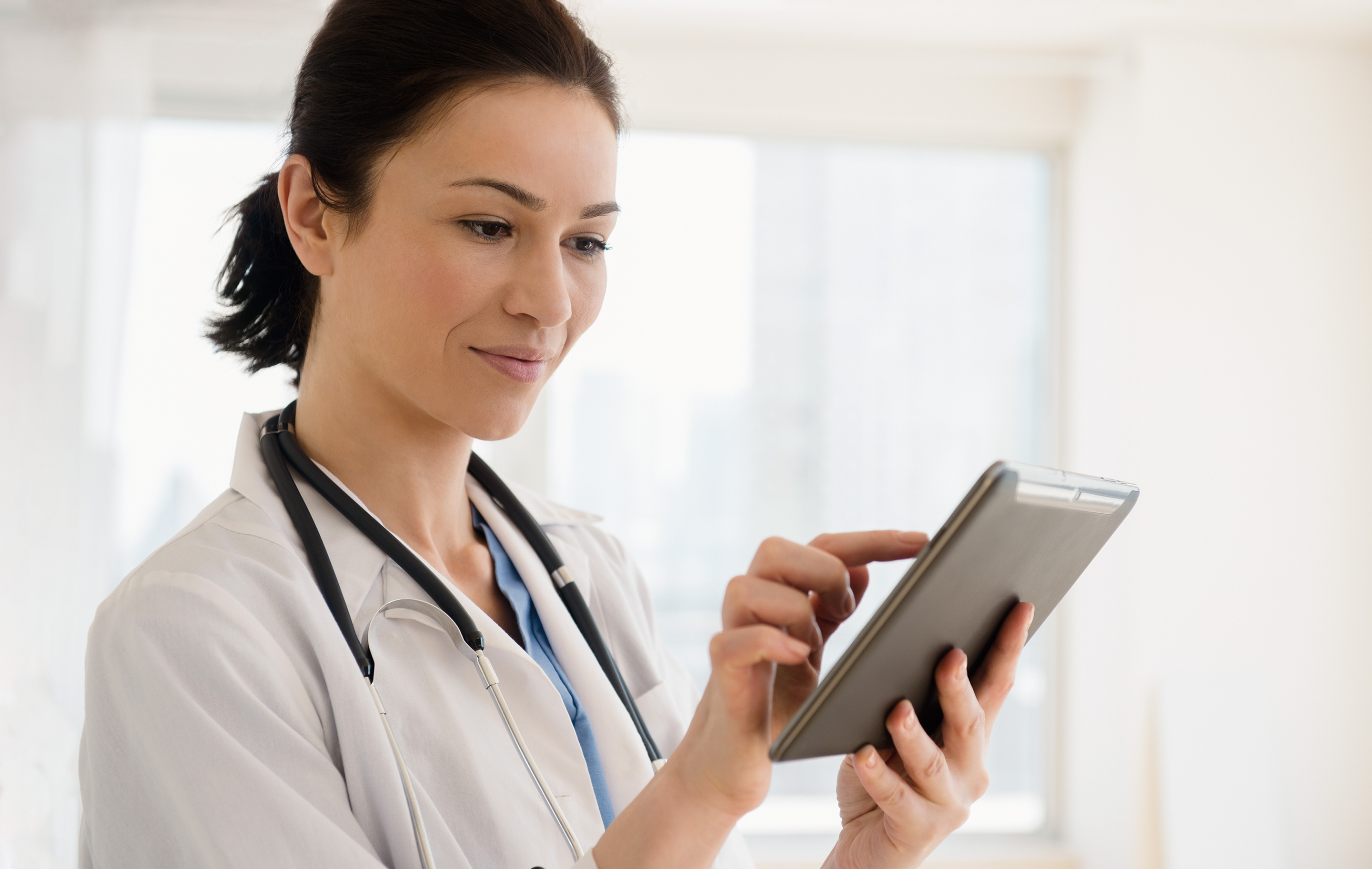 Smiling nurse in white coat with stethoscope uses tablet in bright, clinical setting