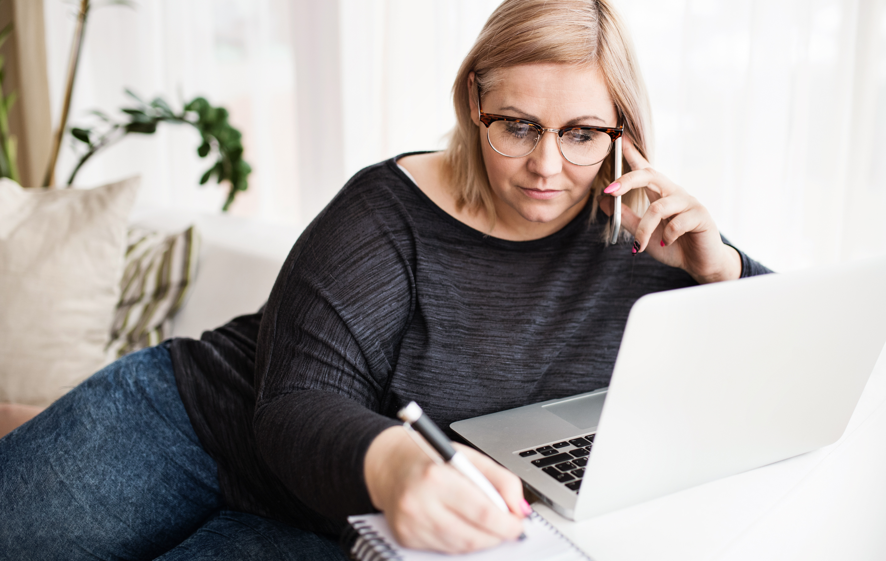 Woman with glasses talks on phone while working on laptop and taking notes at home