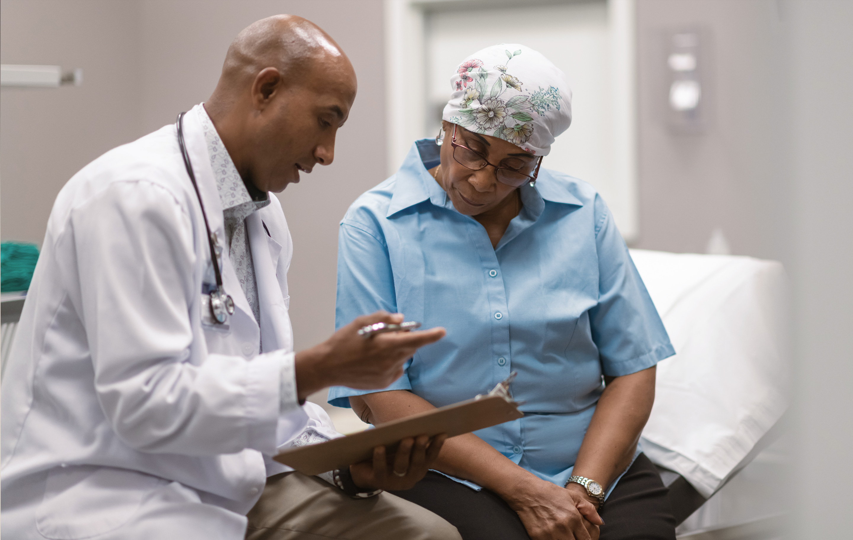Doctor in white coat and stethoscope discusses medical chart with seated patient wearing floral headscarf