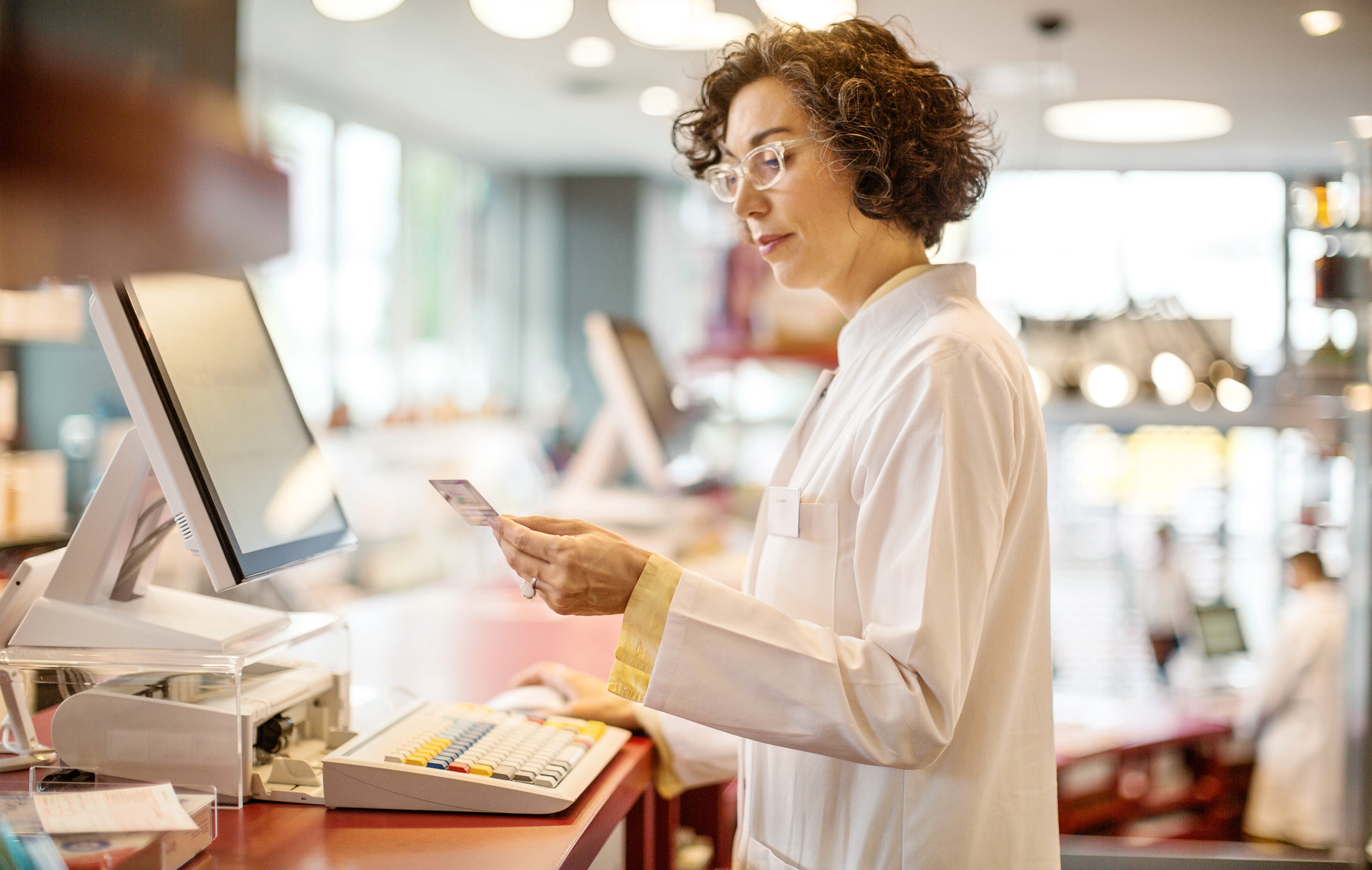 Pharmacist at counter reviewing prescription on tablet with customer in background