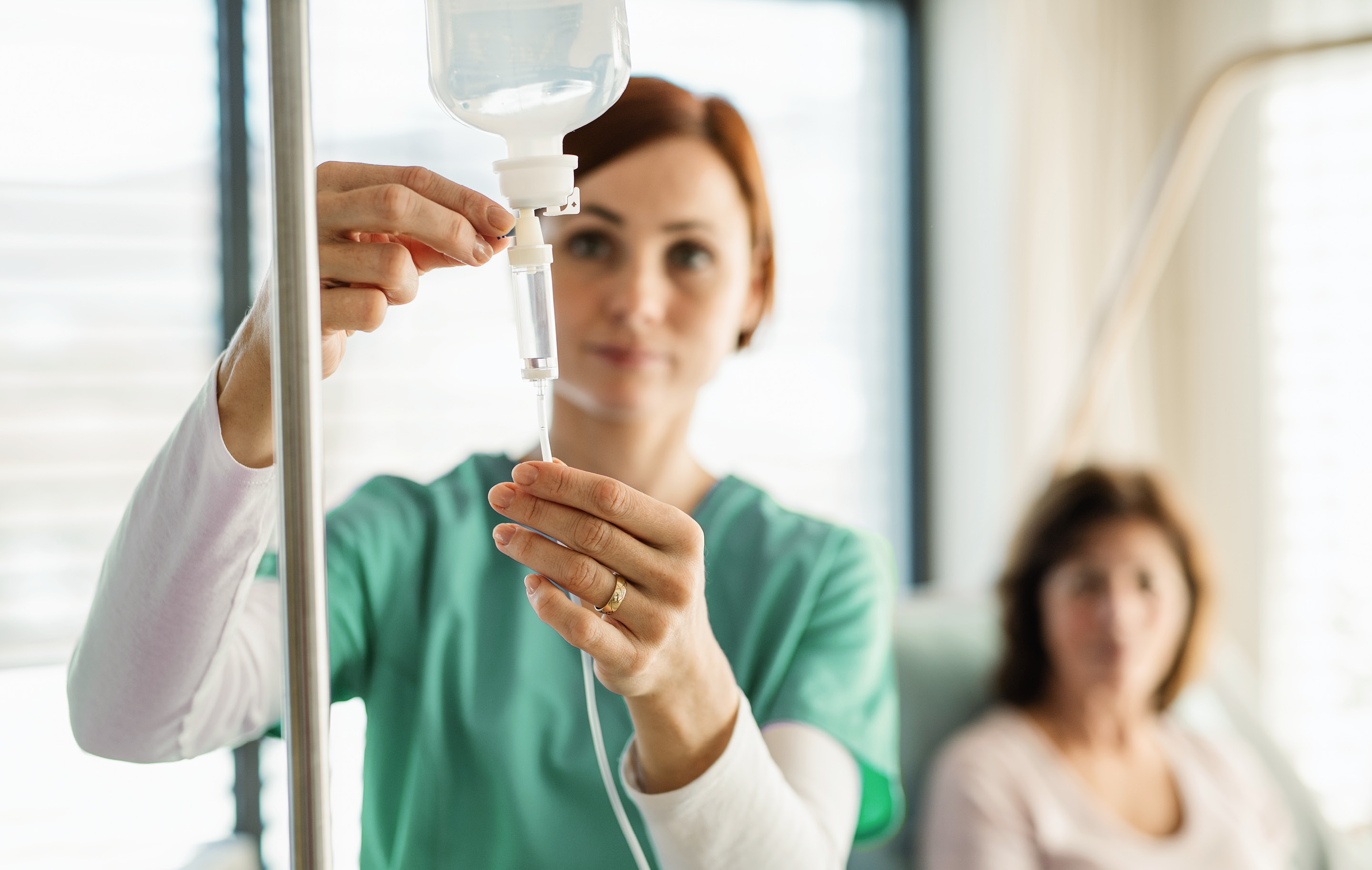Nurse adjusts IV bag in hospital room as patient sits in background