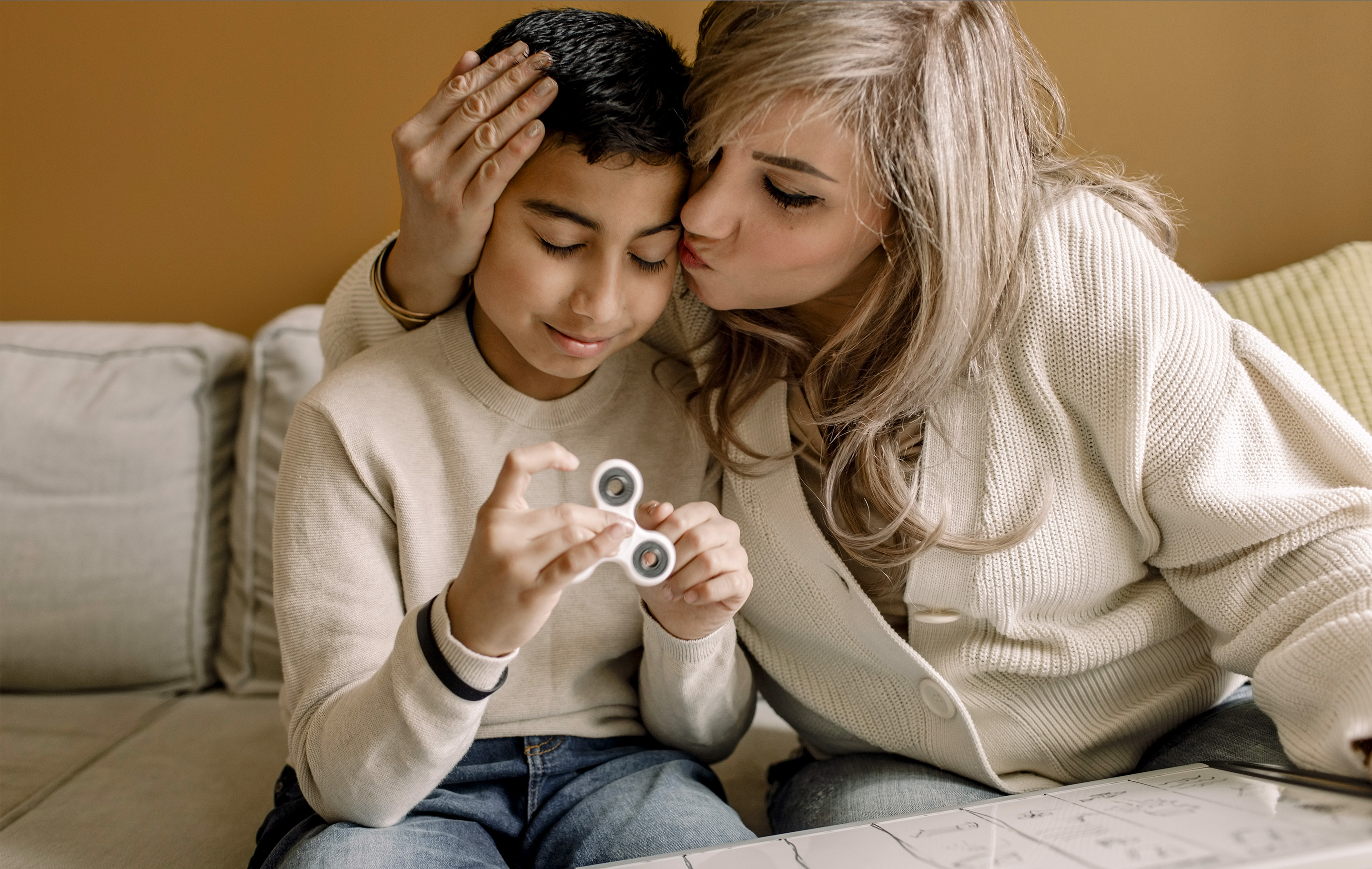 A woman kisses a child's forehead as he holds a fidget spinner on a couch