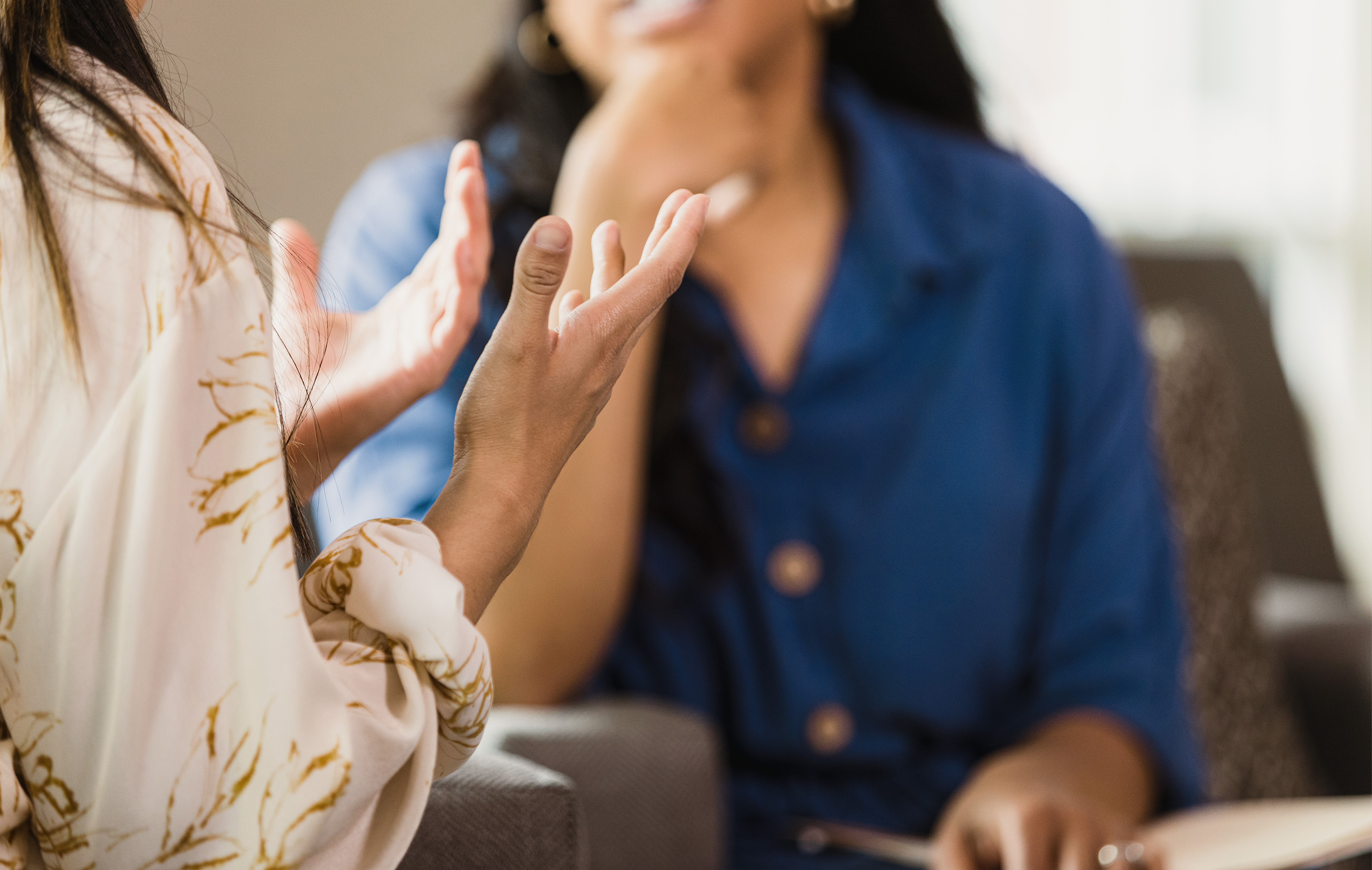 Two women in a softly lit room, one gesturing while seated, the other facing away, suggesting conversation or therapy.
