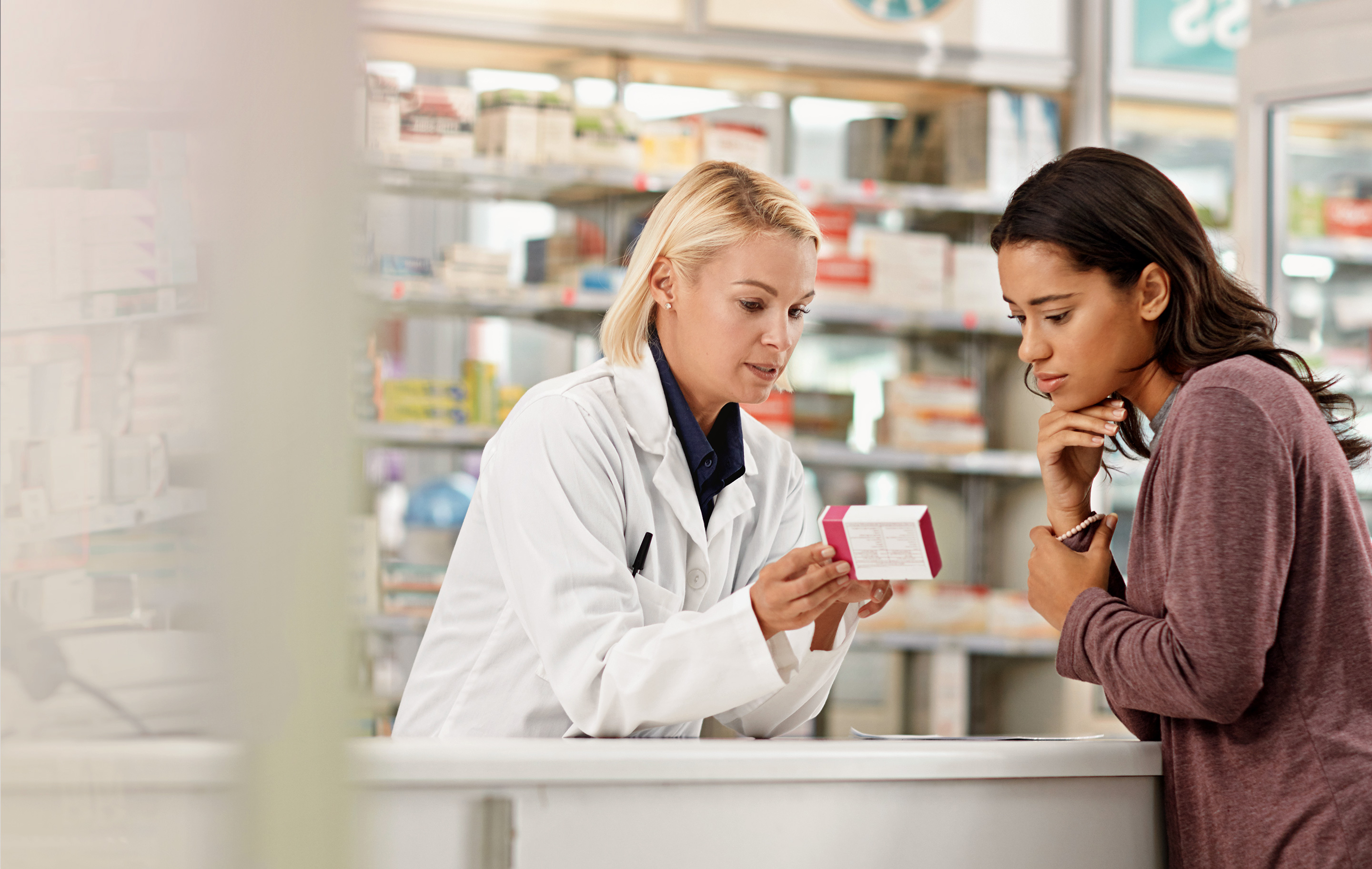 Pharmacist in white coat shows medication to customer at counter in brightly lit pharmacy