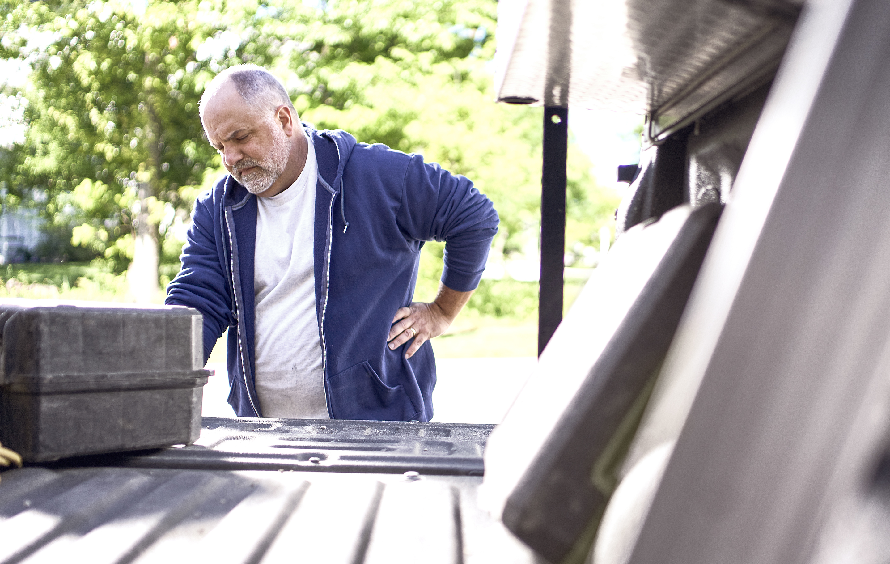Man with beard and blue hoodie leans on truck bed, looking at toolbox in bright outdoor light
