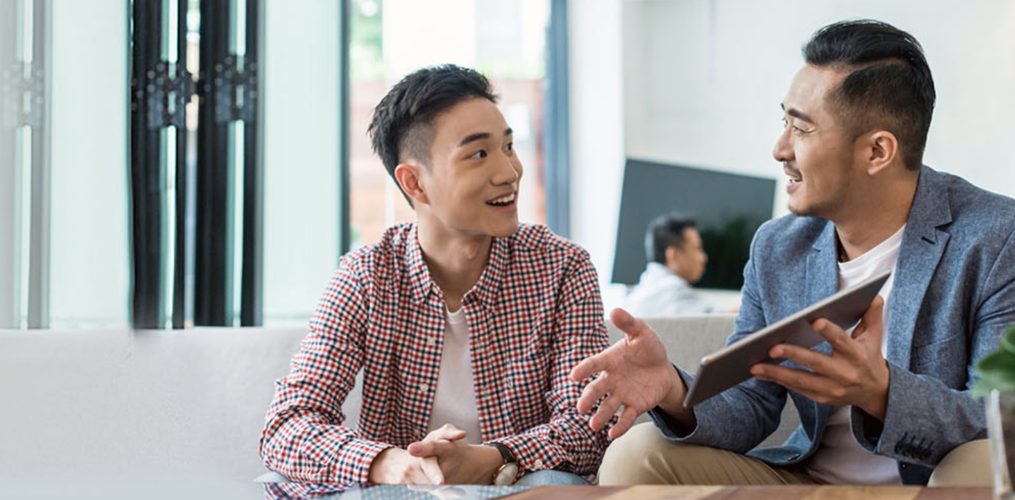 Two men facing each other have conversation. Man on right holds tablet.