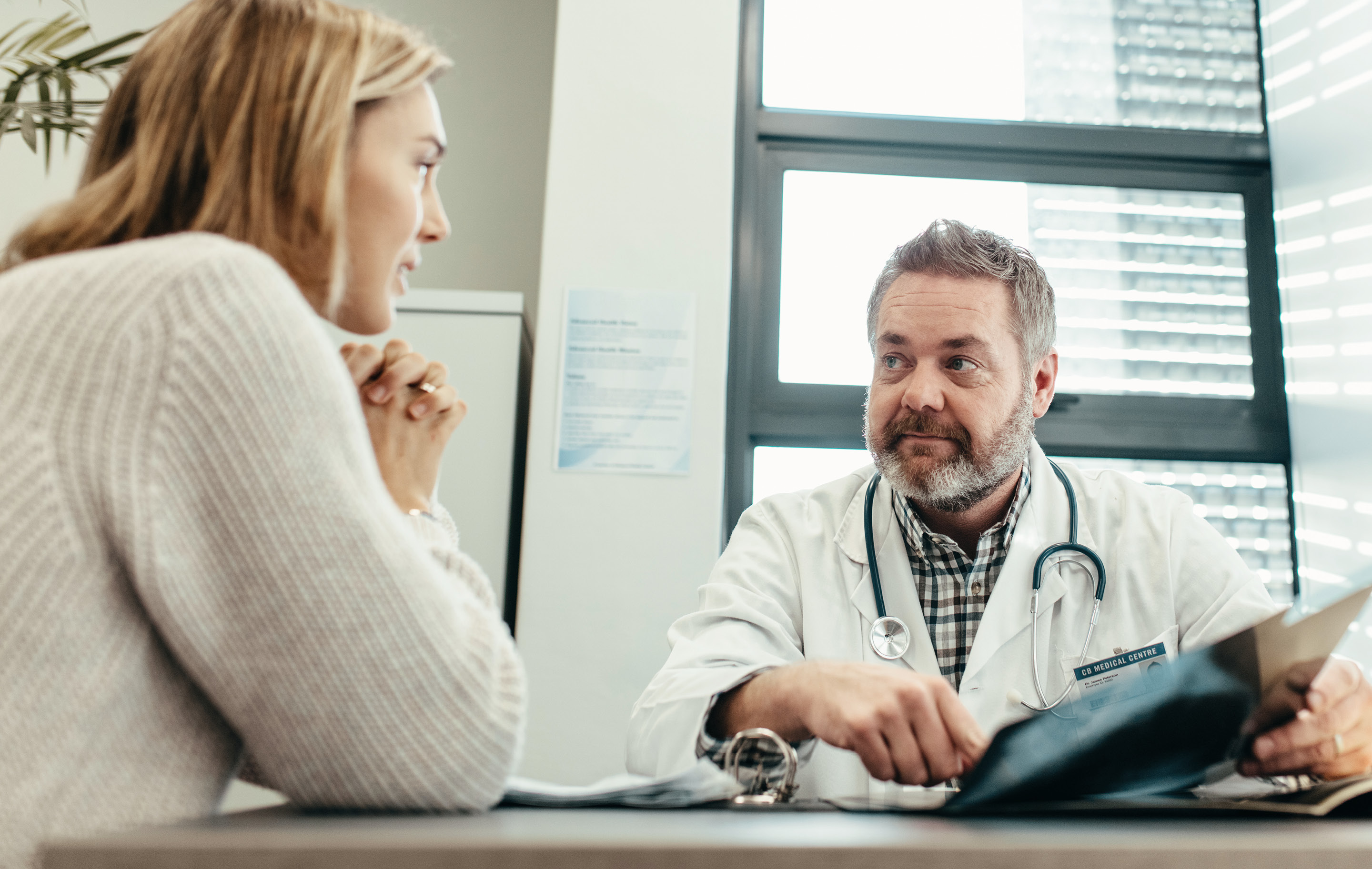 Woman in sweater speaks with male doctor in white coat using tablet at desk in bright clinic room
