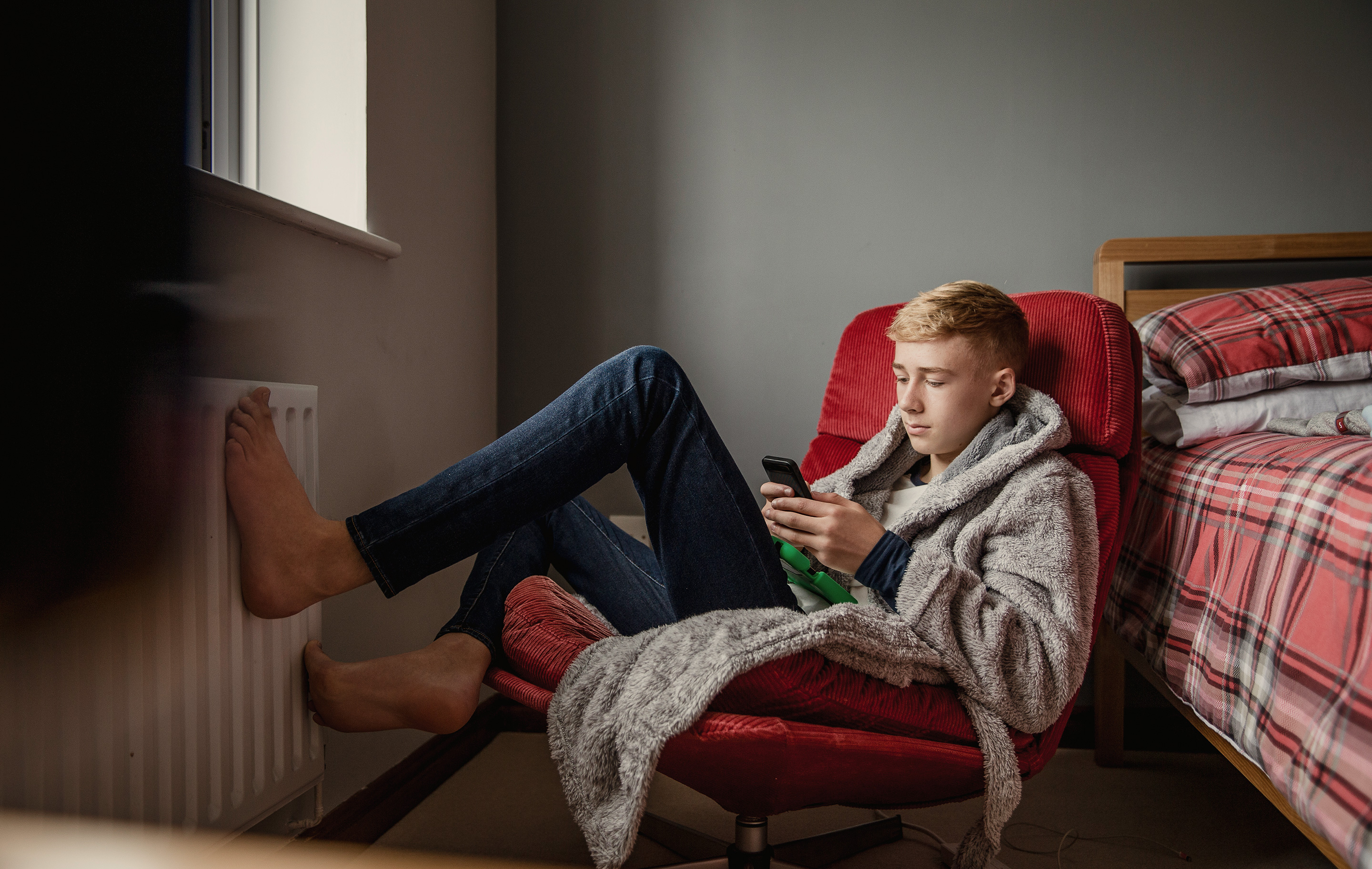Young man in cozy robe sits in red chair by window, feet resting on radiator, phone in hand