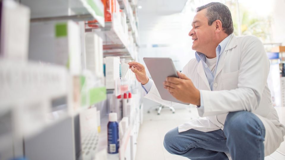 Smiling man in white lab coat crouches in pharmacy aisle holding tablet, reviewing products on shelves