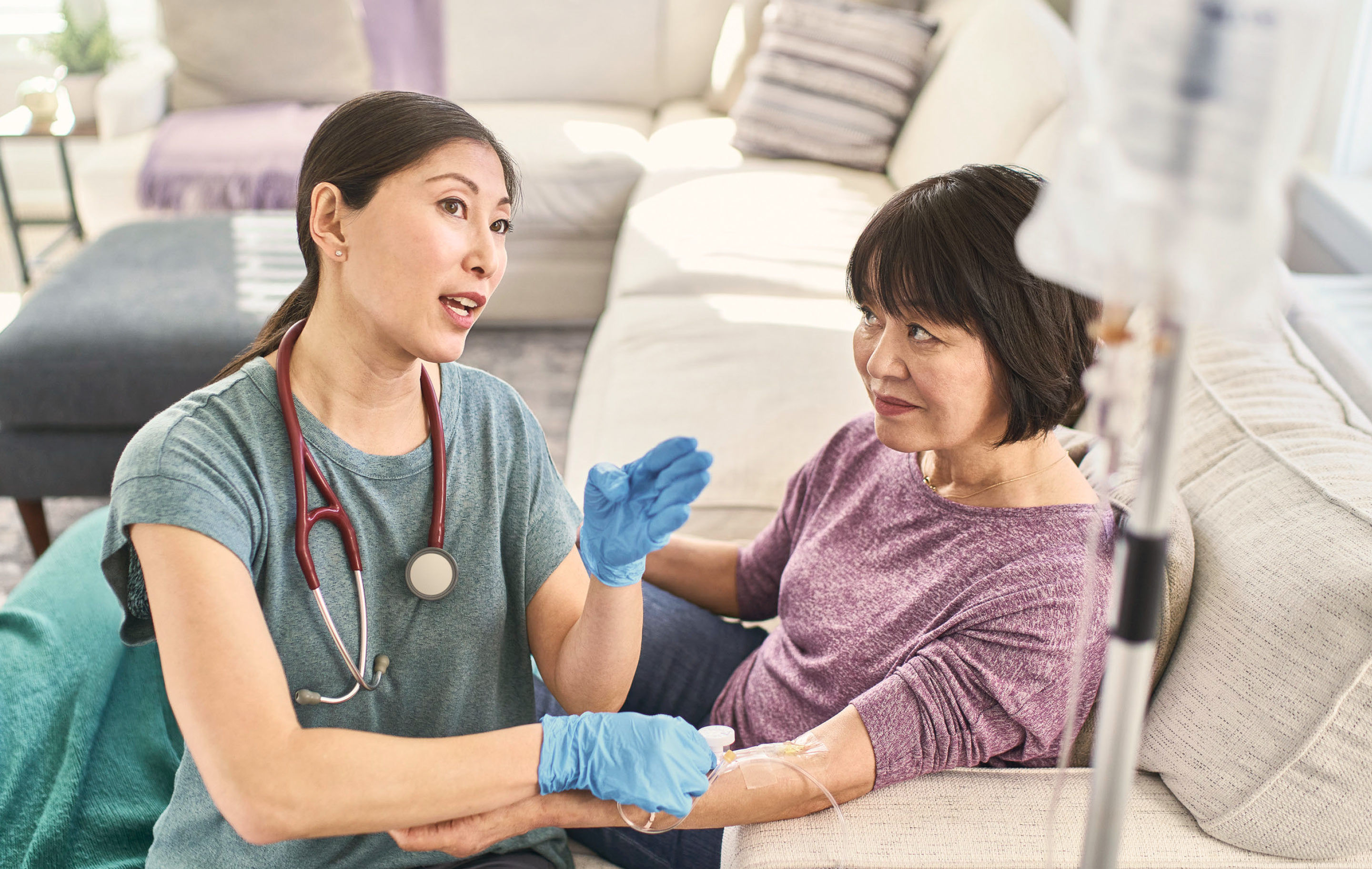 Nurse wearing gloves checks IV line on patient seated in living room sofa with medical equipment nearby