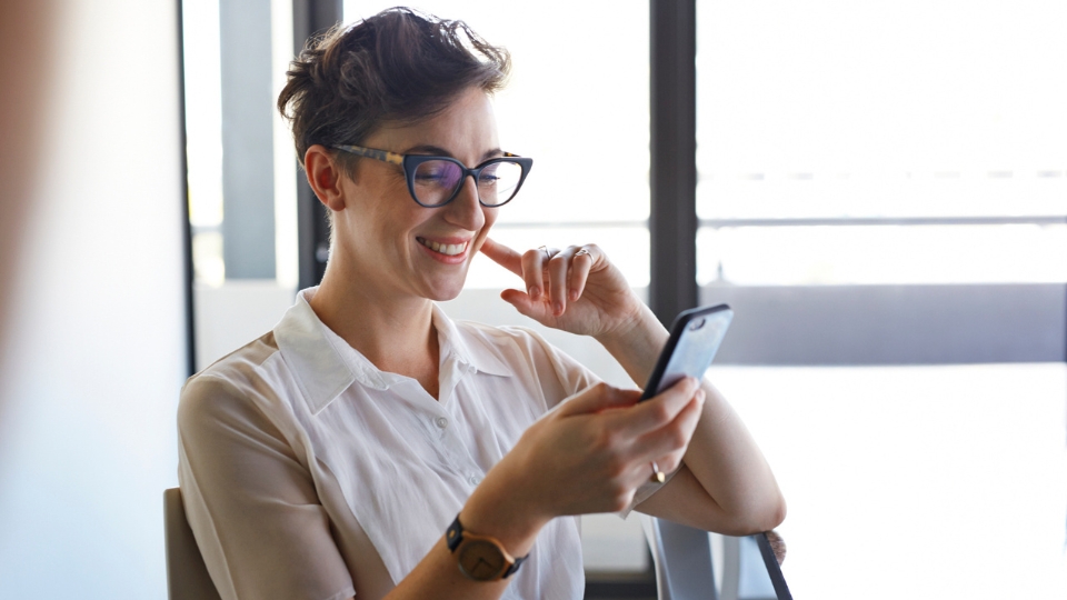 Smiling woman with glasses uses smartphone near window, wearing white blouse and watch