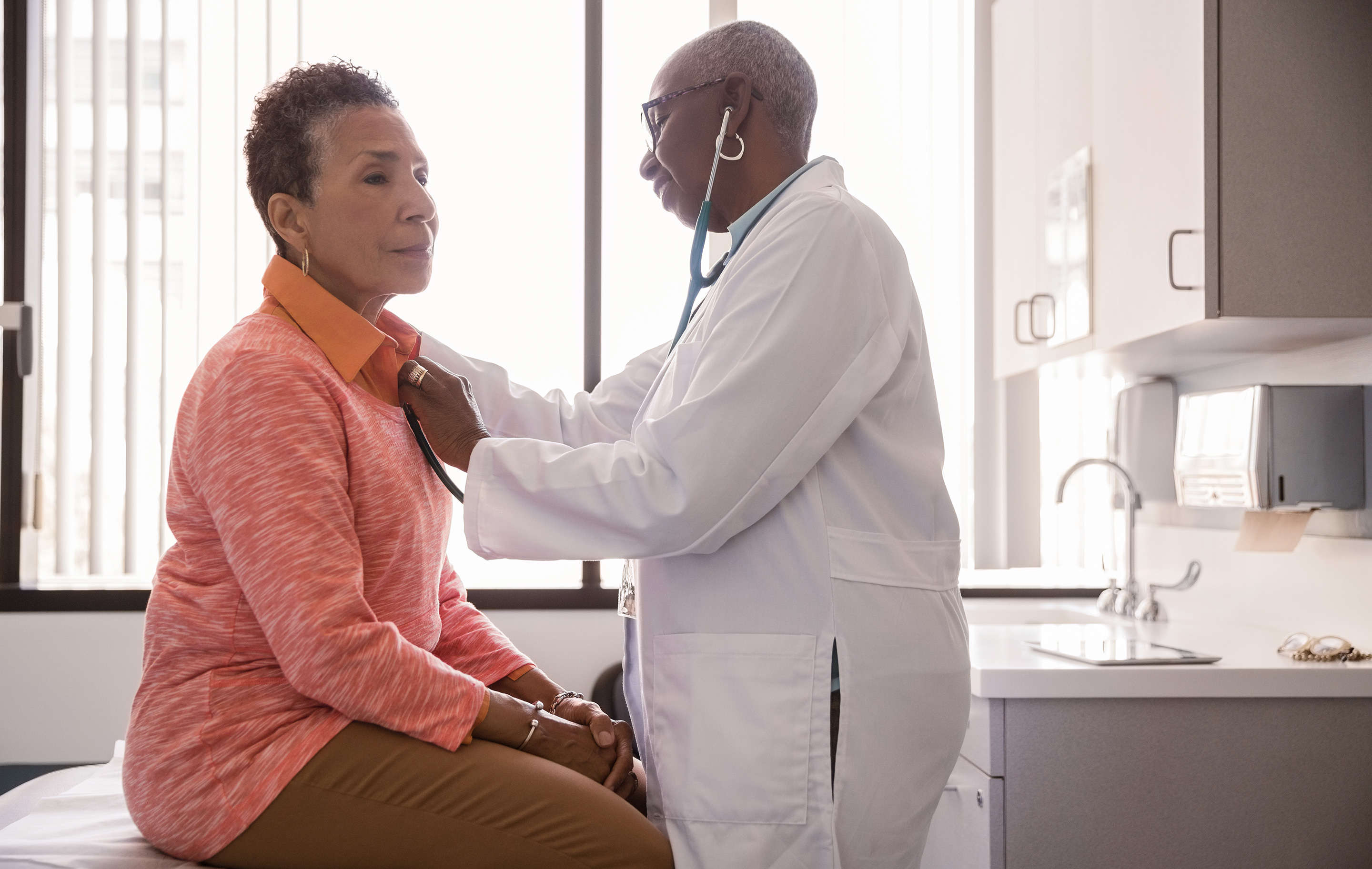 Doctor uses stethoscope on patient seated on exam table in bright clinic room