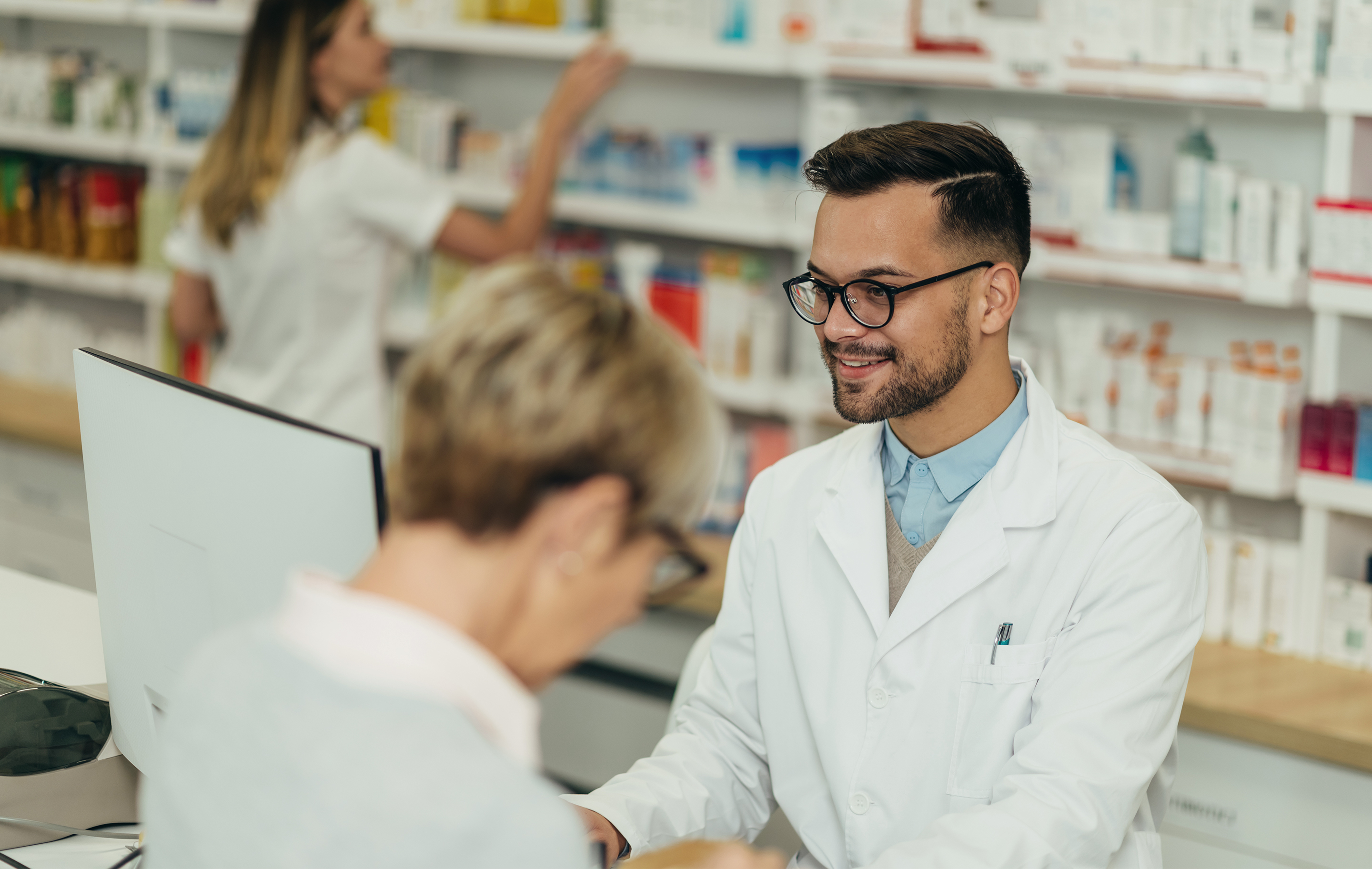 Pharmacist in white coat smiles while assisting customer at counter