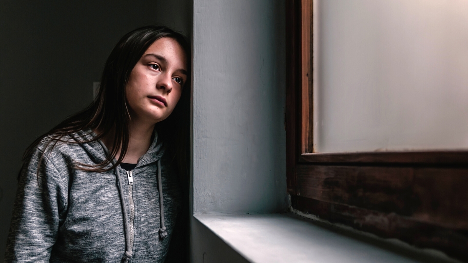 A young woman leans against a window sill in dim light, gazing outward with a pensive expression.