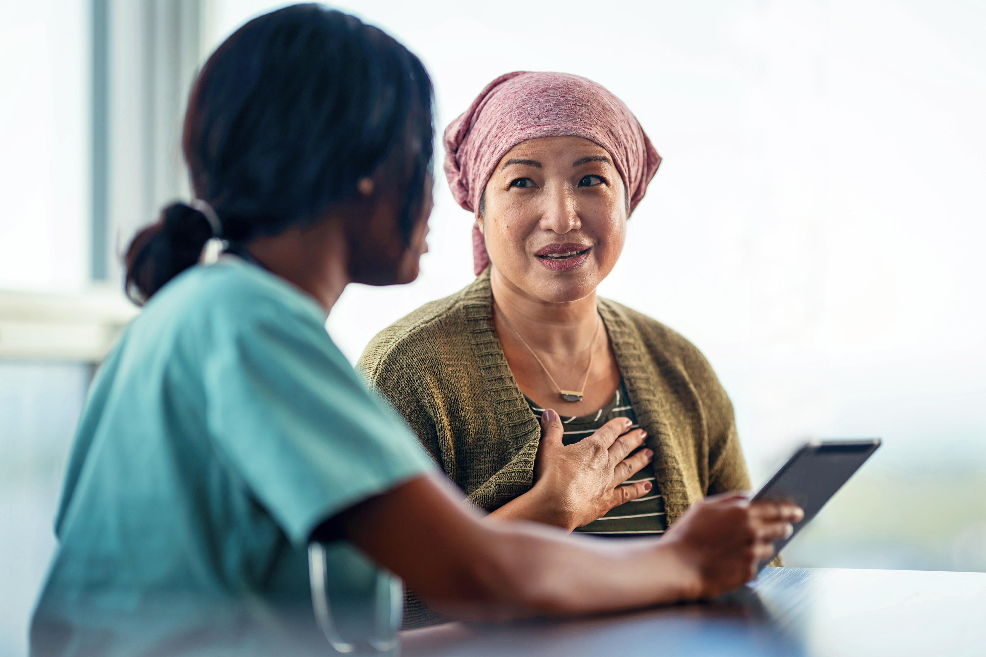 A medical professional who is wearing scrubs holds a tablet and speaks with a patient