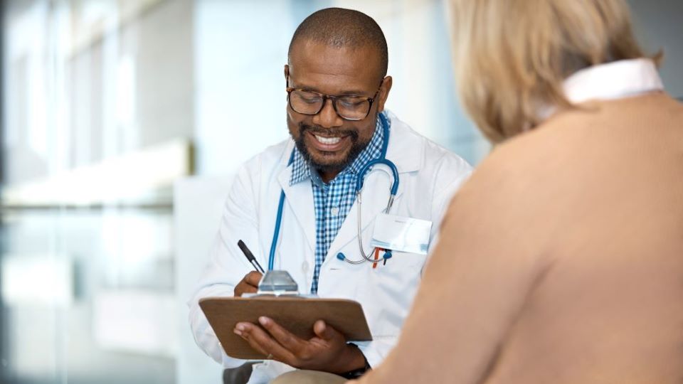 Smiling doctor in white coat uses tablet with stethoscope, consulting with patient in bright hospital hallway