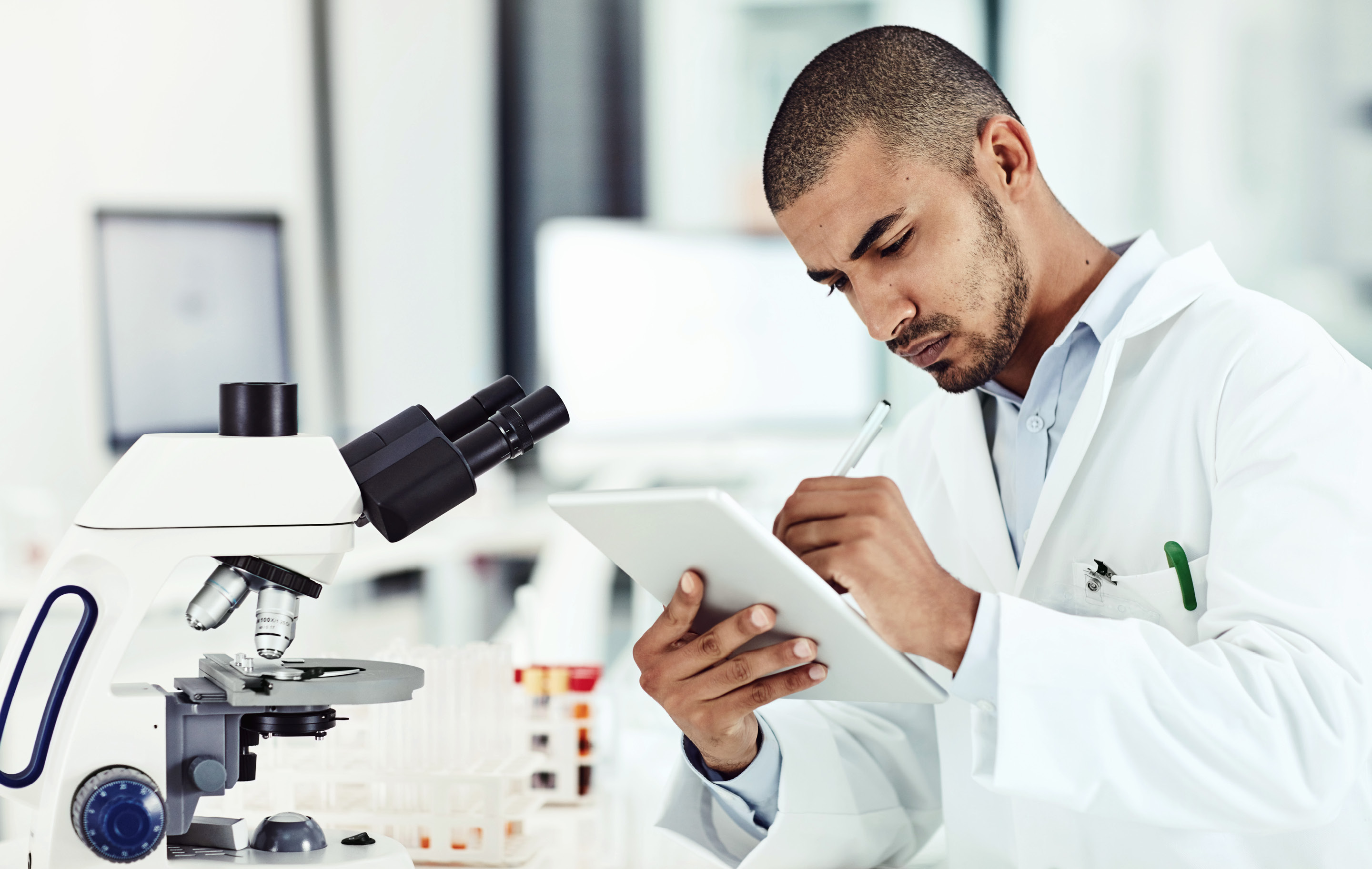 Scientist in lab coat examines sample under microscope while noting observations on tablet.