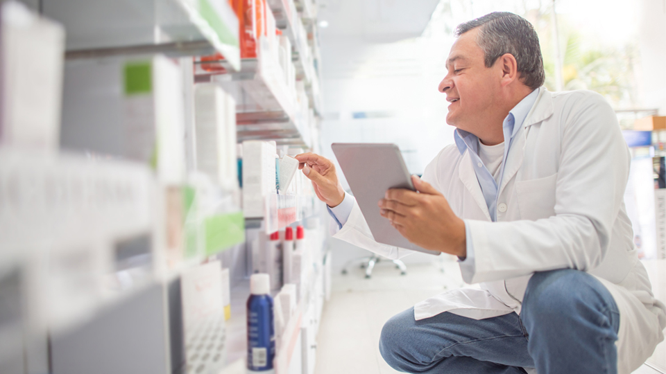 Doctor crouches in pharmacy aisle holding tablet, reviewing product labels with focused expression.