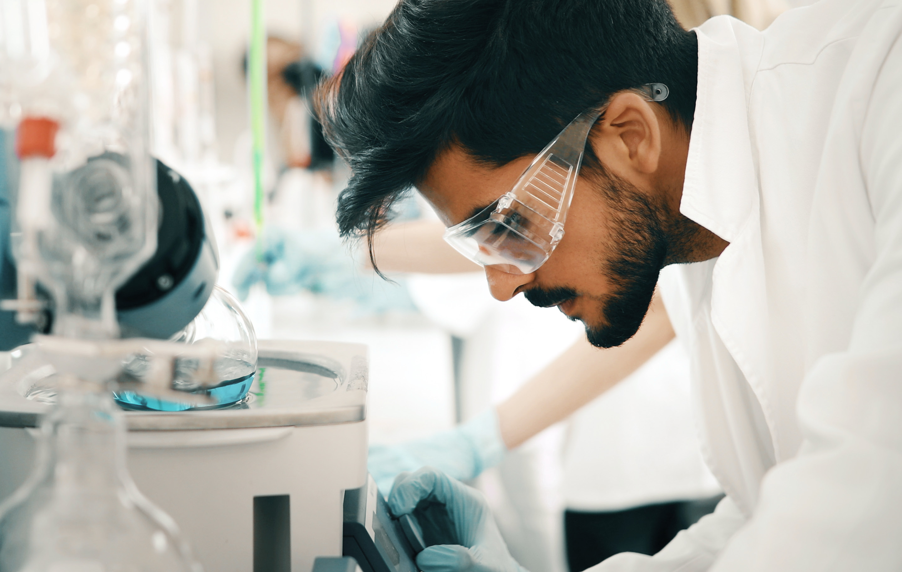 Scientist in lab coat and goggles examines sample under microscope in bright, clinical setting.