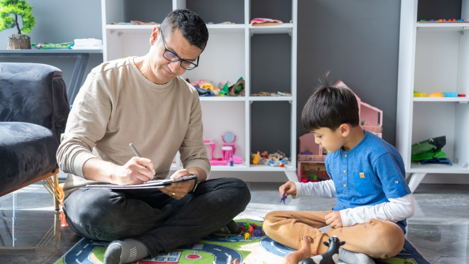 Man and child sit on floor in cozy room with toys and books nearby.