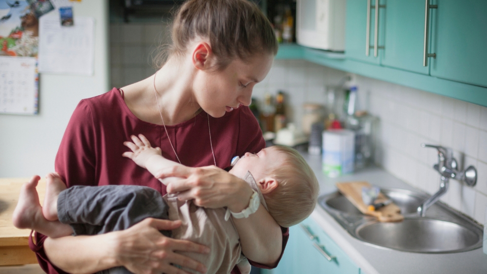 Mother cradles infant in kitchen, gently holding baby close with soft, caring expression.