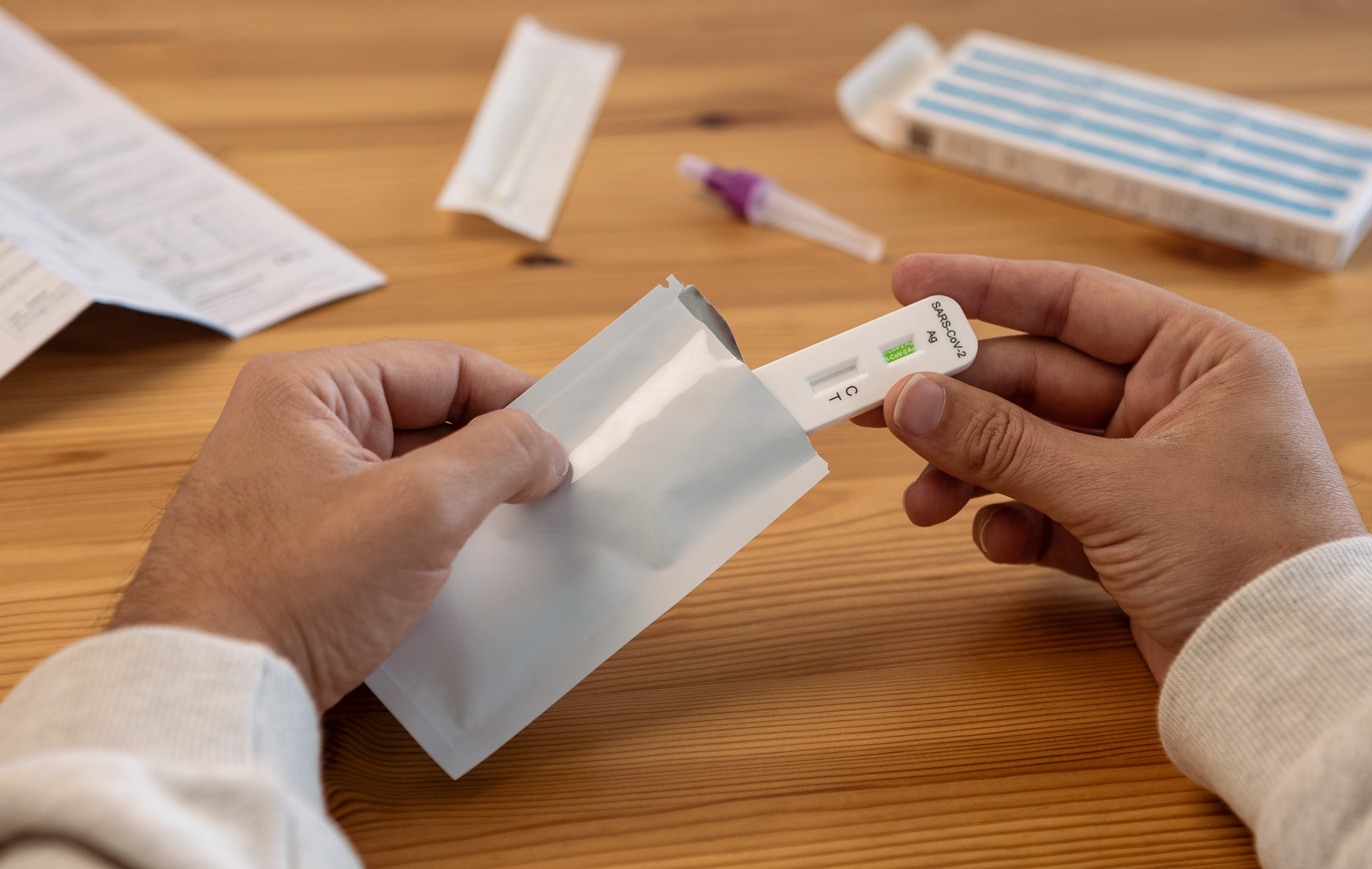 Hands hold a COVID-19 test kit on a wooden table with other medical supplies nearby.