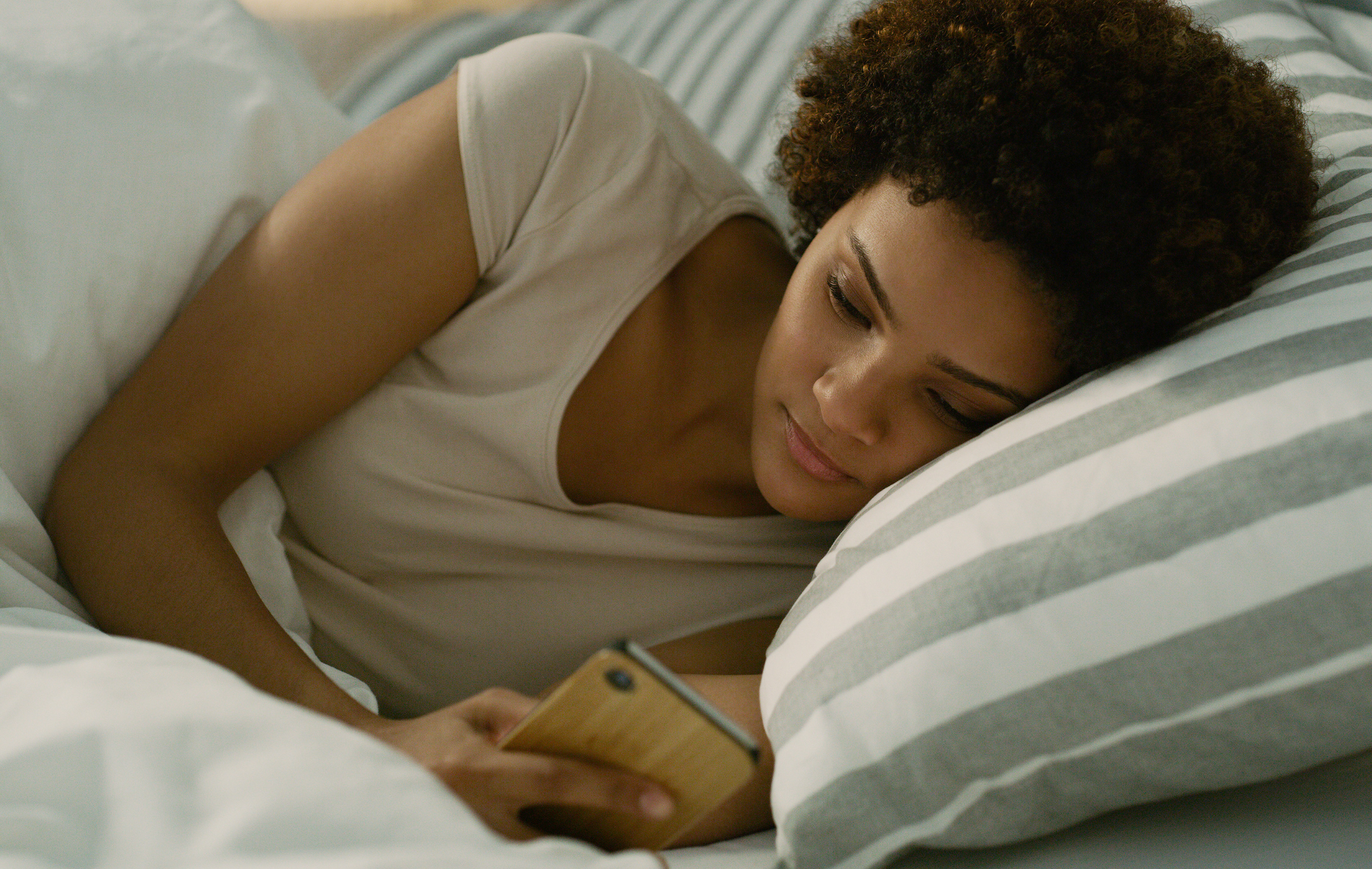 Woman lying in bed with phone, head on pillow, soft lighting, relaxed expression.