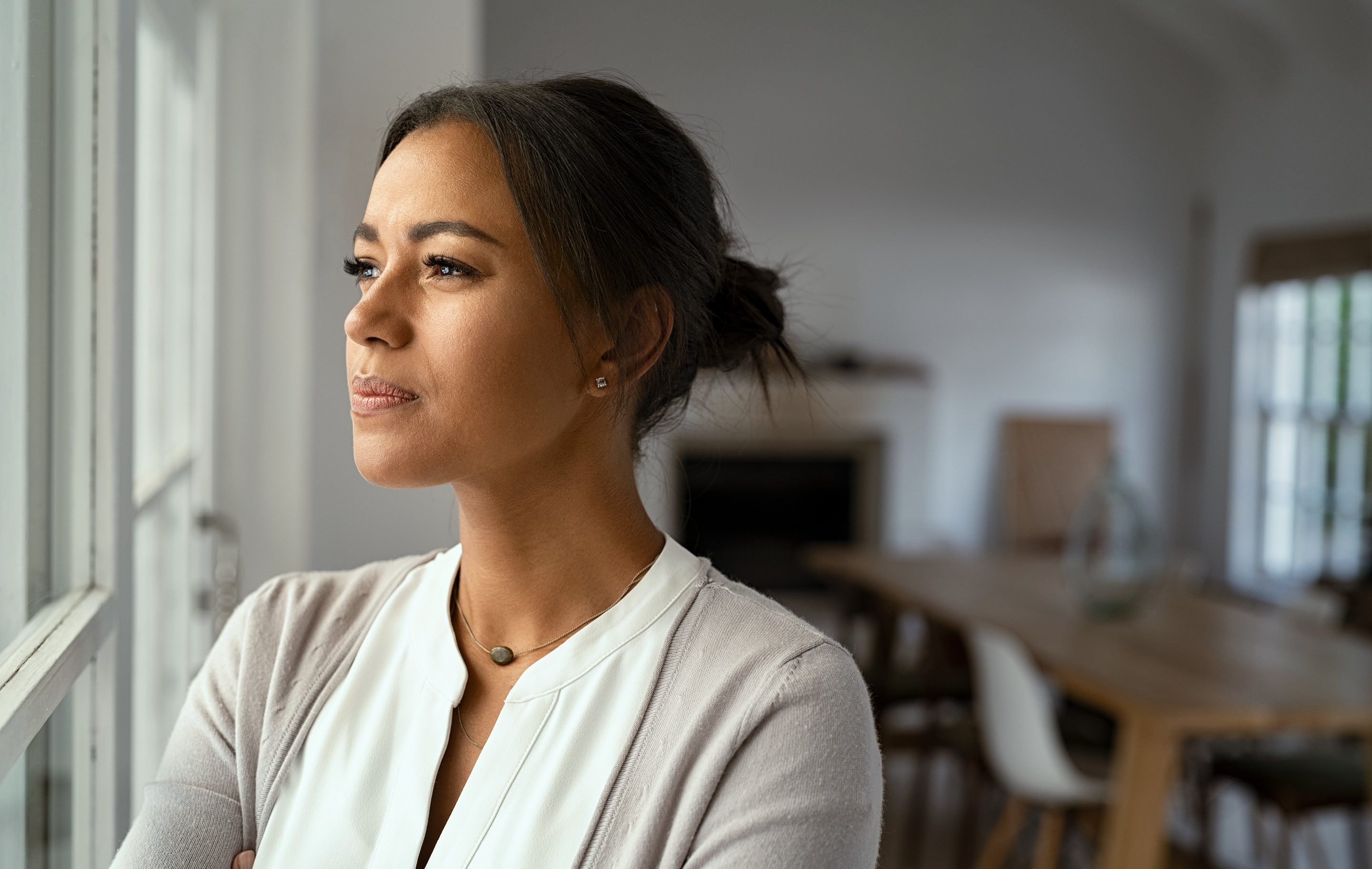 Woman stands by window in bright room looking outward with calm expression.