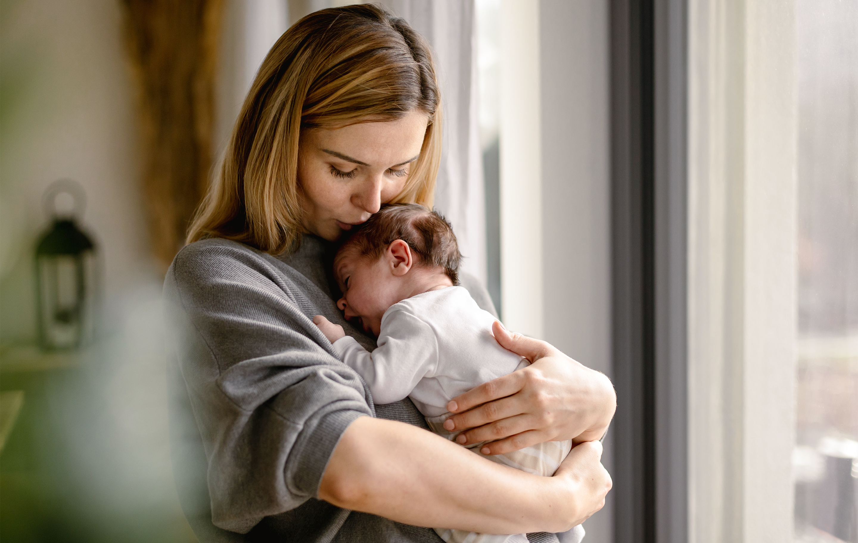 A woman cradles a sleeping infant close to a window, bathed in soft natural light.