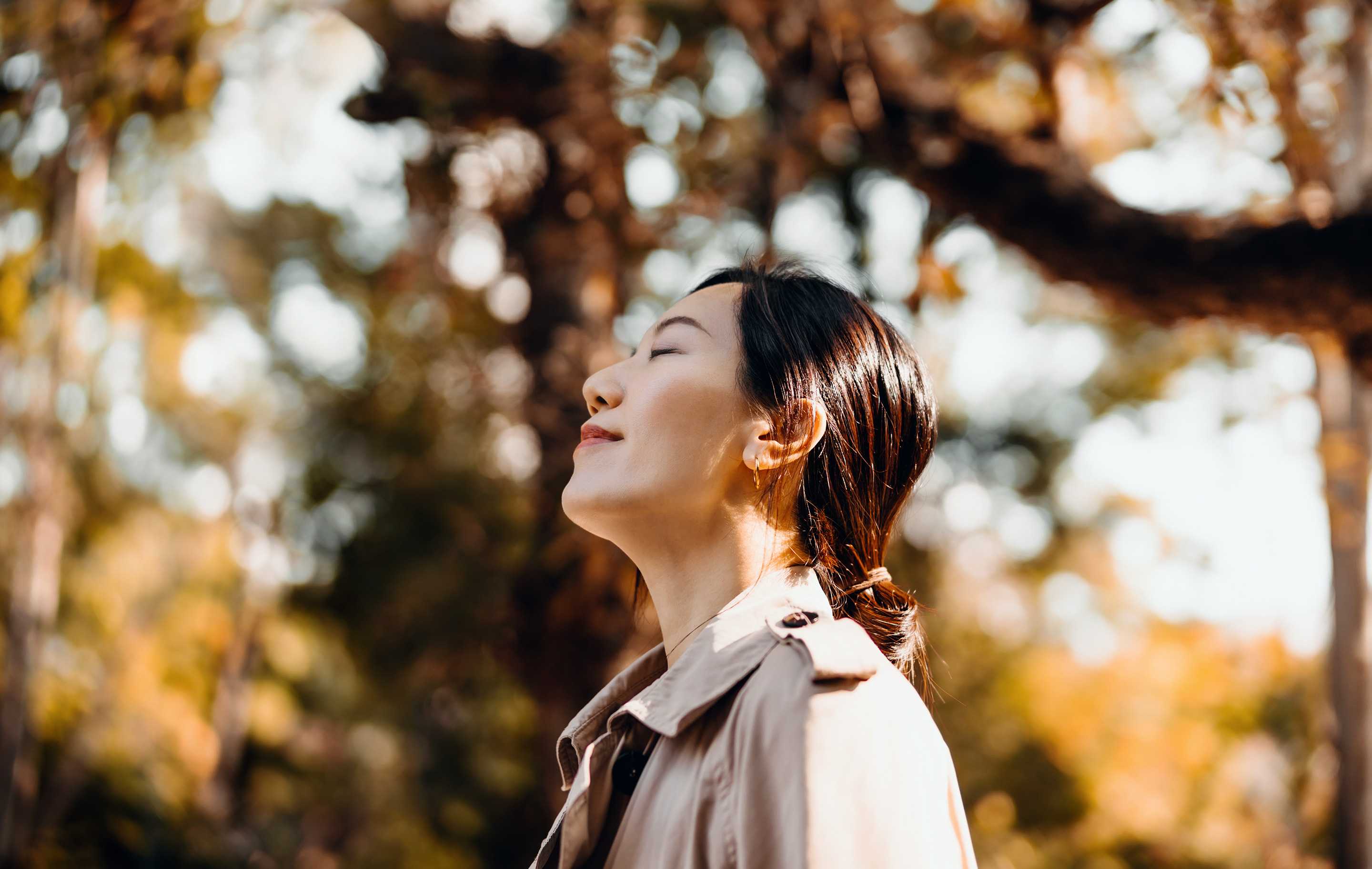 Woman in trench coat closes eyes, breathing deeply, sun-dappled autumn trees behind her.