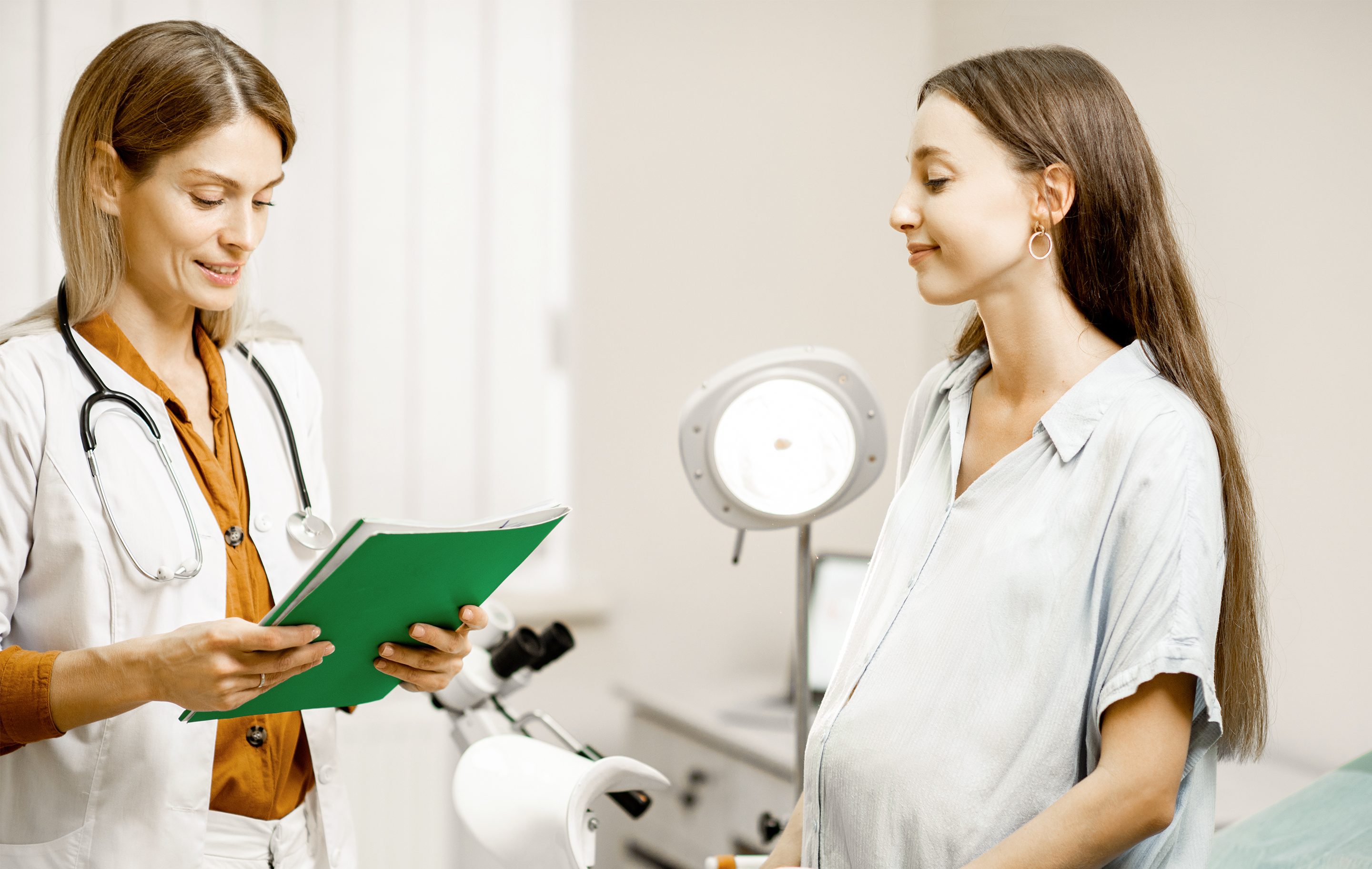 Doctor consults with pregnant patient in clinical setting holding green folder with bright lighting.