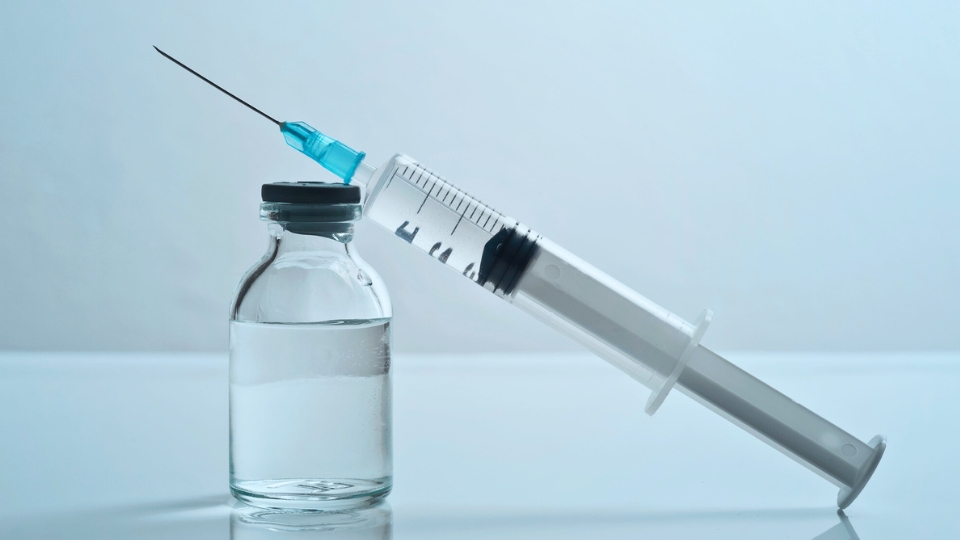 Syringe with needle resting beside a glass vial of clear liquid on a light surface.
