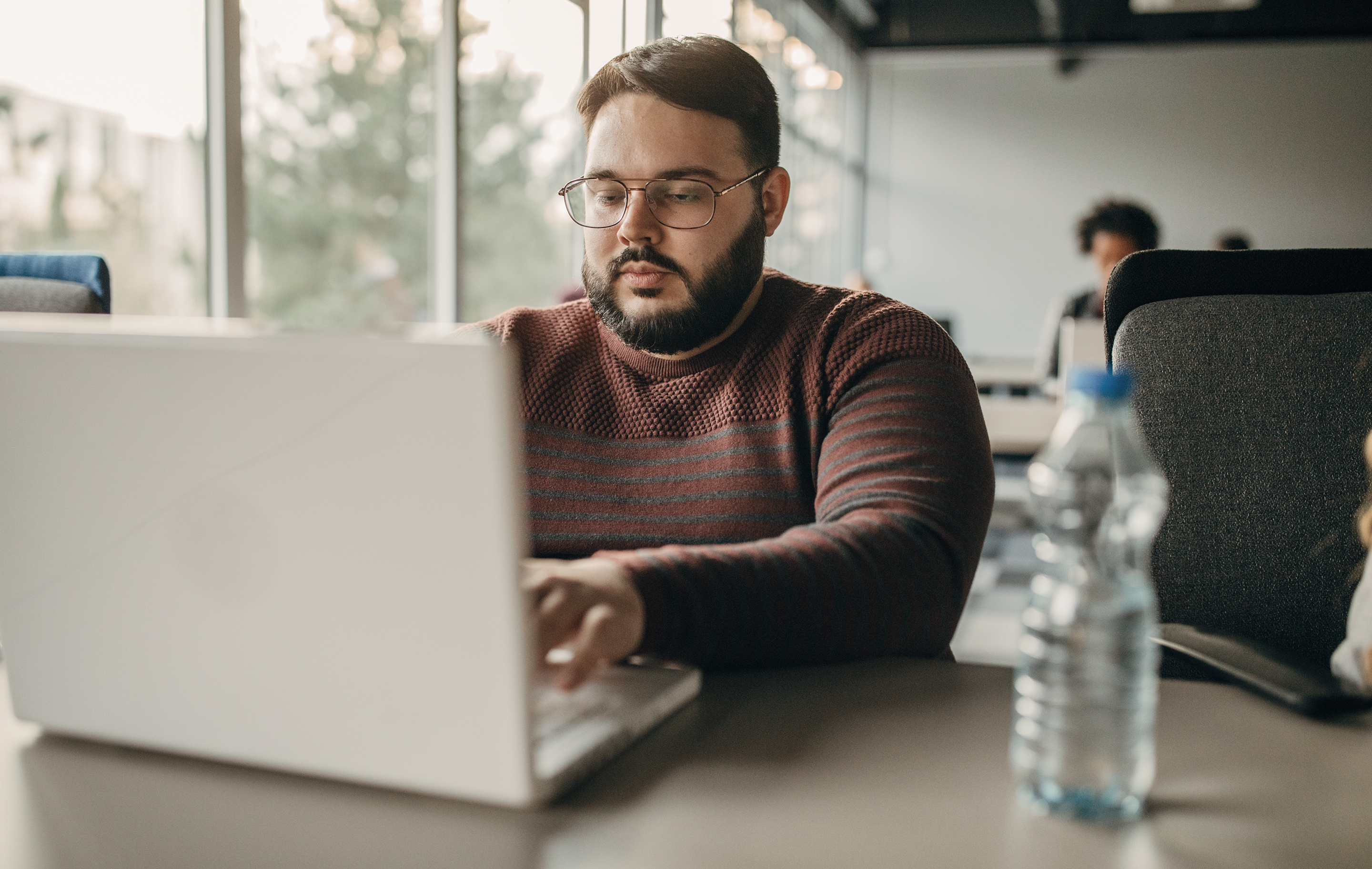 Man with glasses works on laptop at desk with water bottle nearby in bright office space