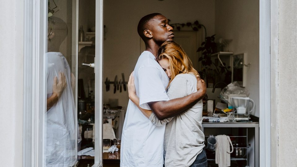 A man and woman embrace in a softly lit kitchen, conveying quiet comfort.