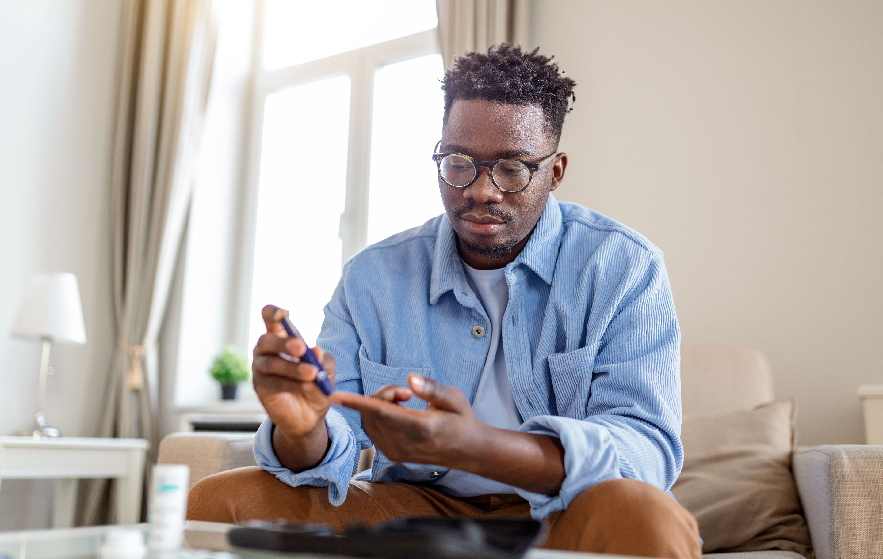 Man with glasses checks blood sugar levels while seated in a brightly lit home setting.