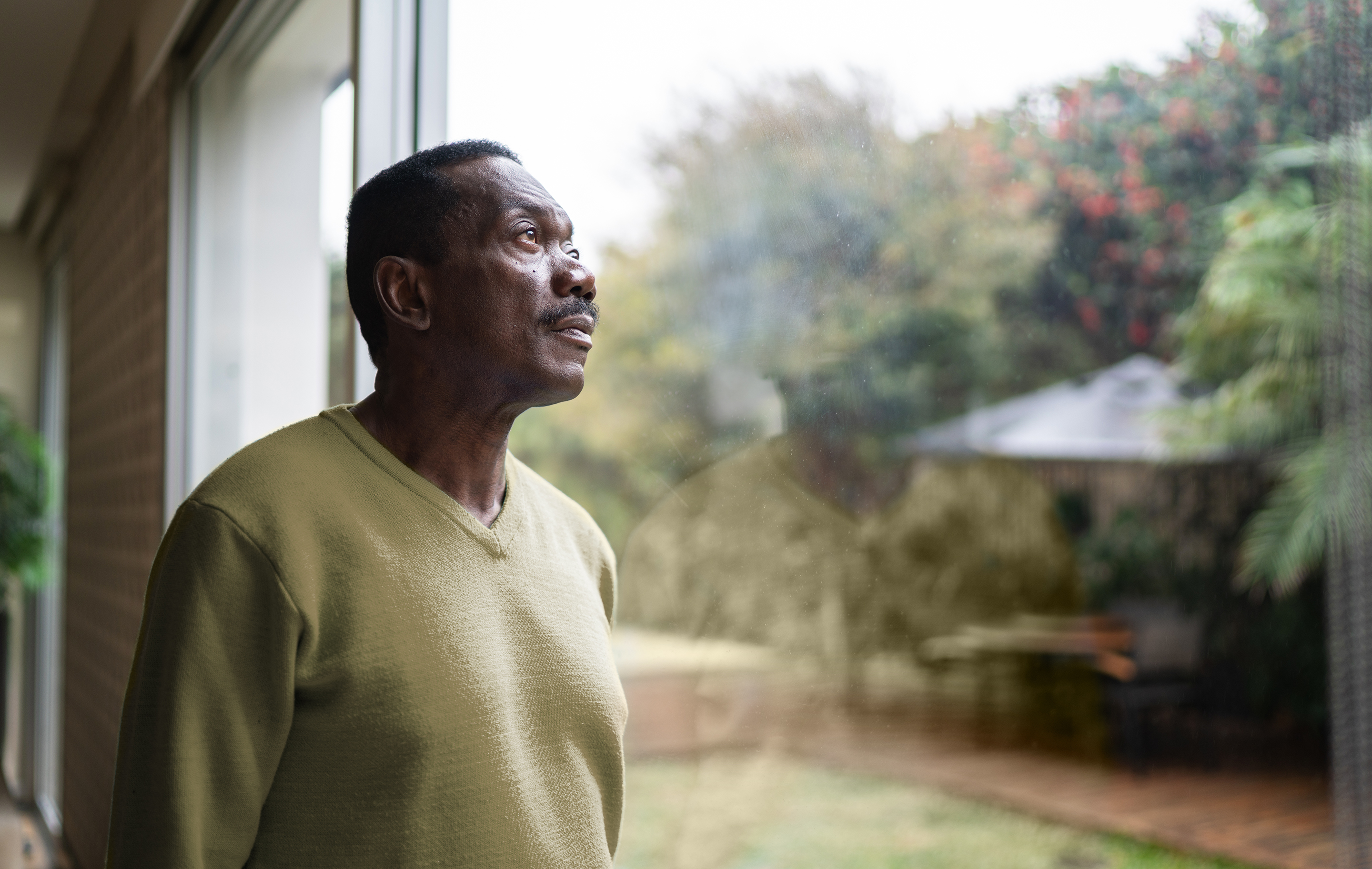 Man in green sweater gazes out a window at a blurred garden scene.