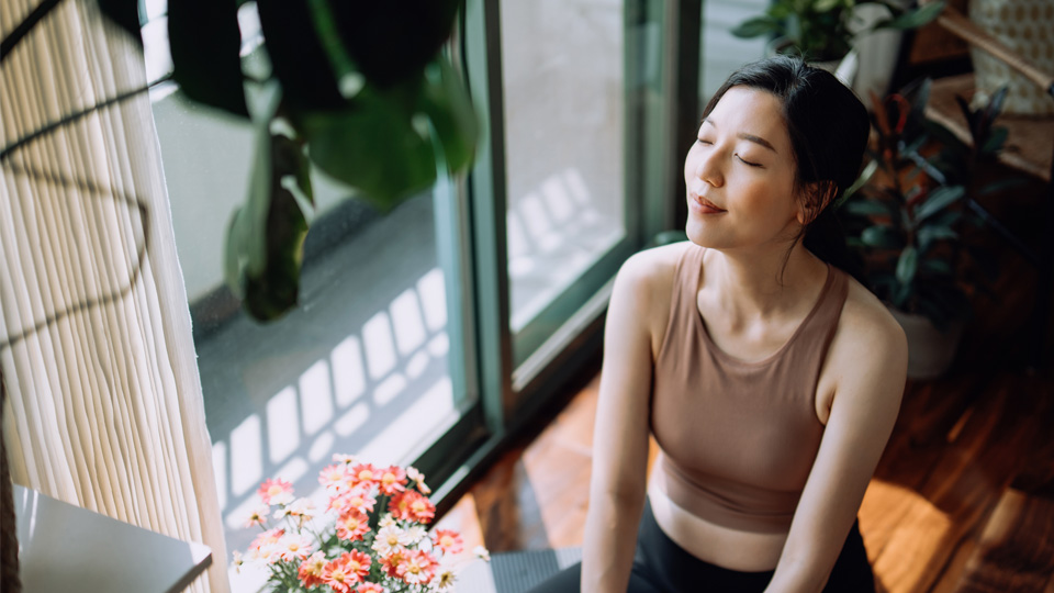 Woman smiles softly, bathed in sunlight near window with plants and flowers nearby.