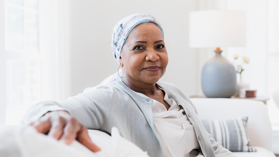 A woman wearing a headscarf sits on a couch in a softly lit room, looking thoughtfully ahead.