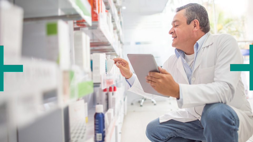 Doctor crouches in pharmacy aisle holding tablet, examining product label
