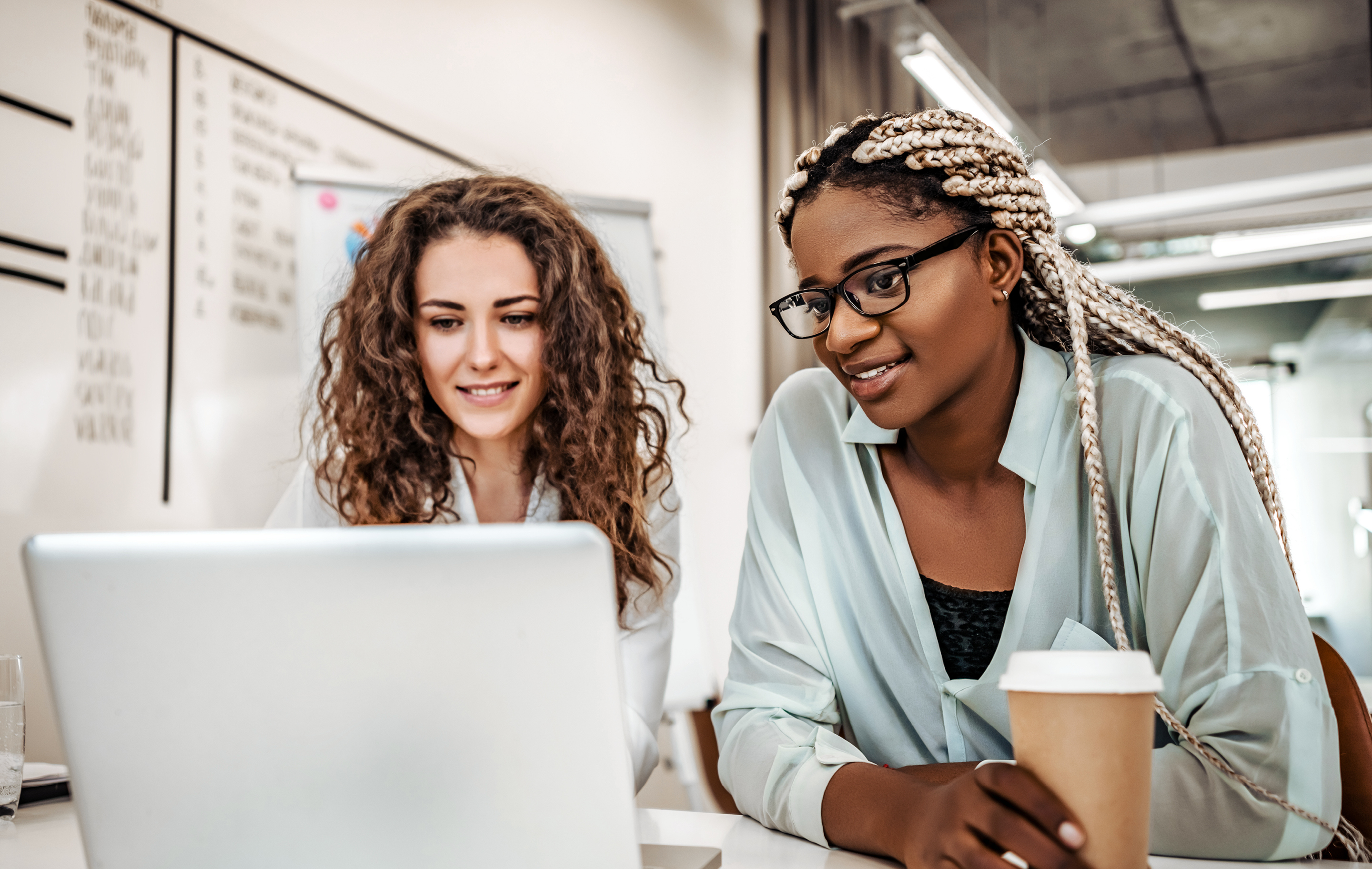 Two colleagues collaborate at a laptop in a bright office, one holding coffee, smiling together.