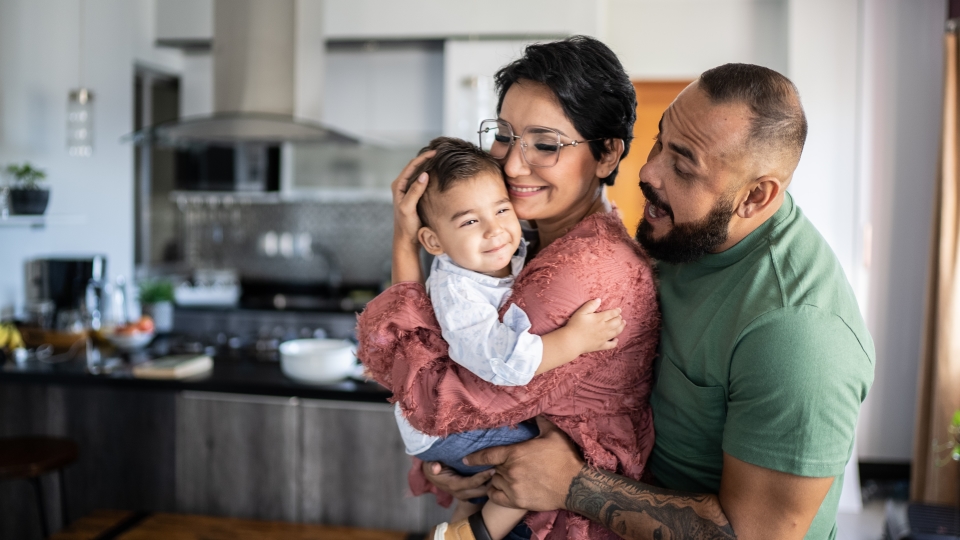 A family of three smile and embrace each other in a bright kitchen setting